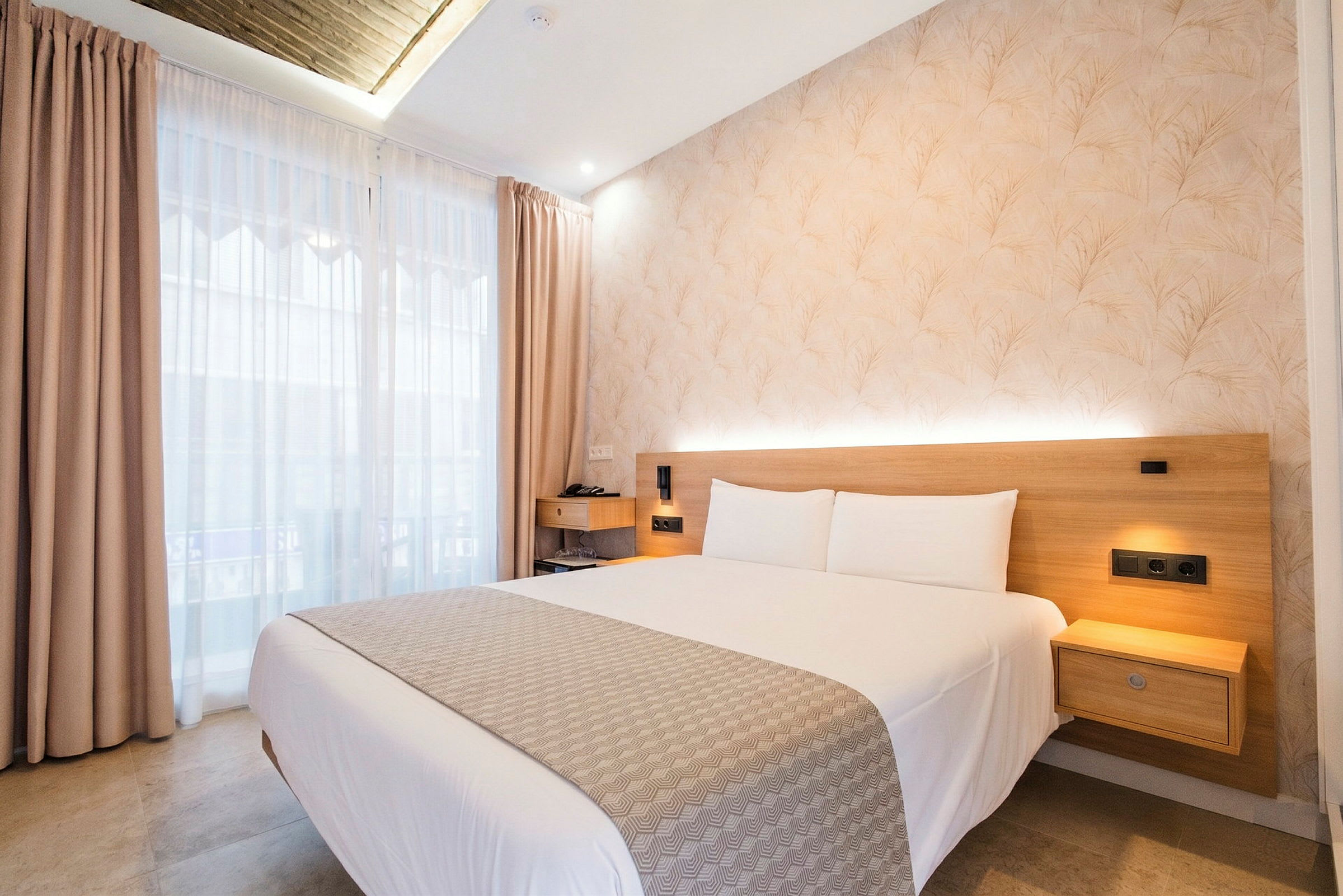 Ett hotellrum med en dubbelsäng, ljust sängöverkast och kuddar. En ljus vägg med träpanel och en liten nattygsbord är synliga. Fönster med ljusa gardiner släpper in dagsljus. (AI-genererad text)