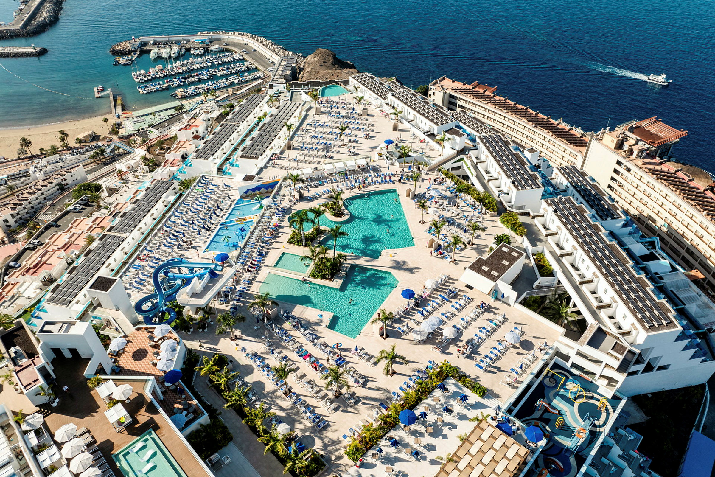 Flygfoto över hotellområde med pooler, solstolar och omgivande byggnader nära havet. (AI-genererad text)