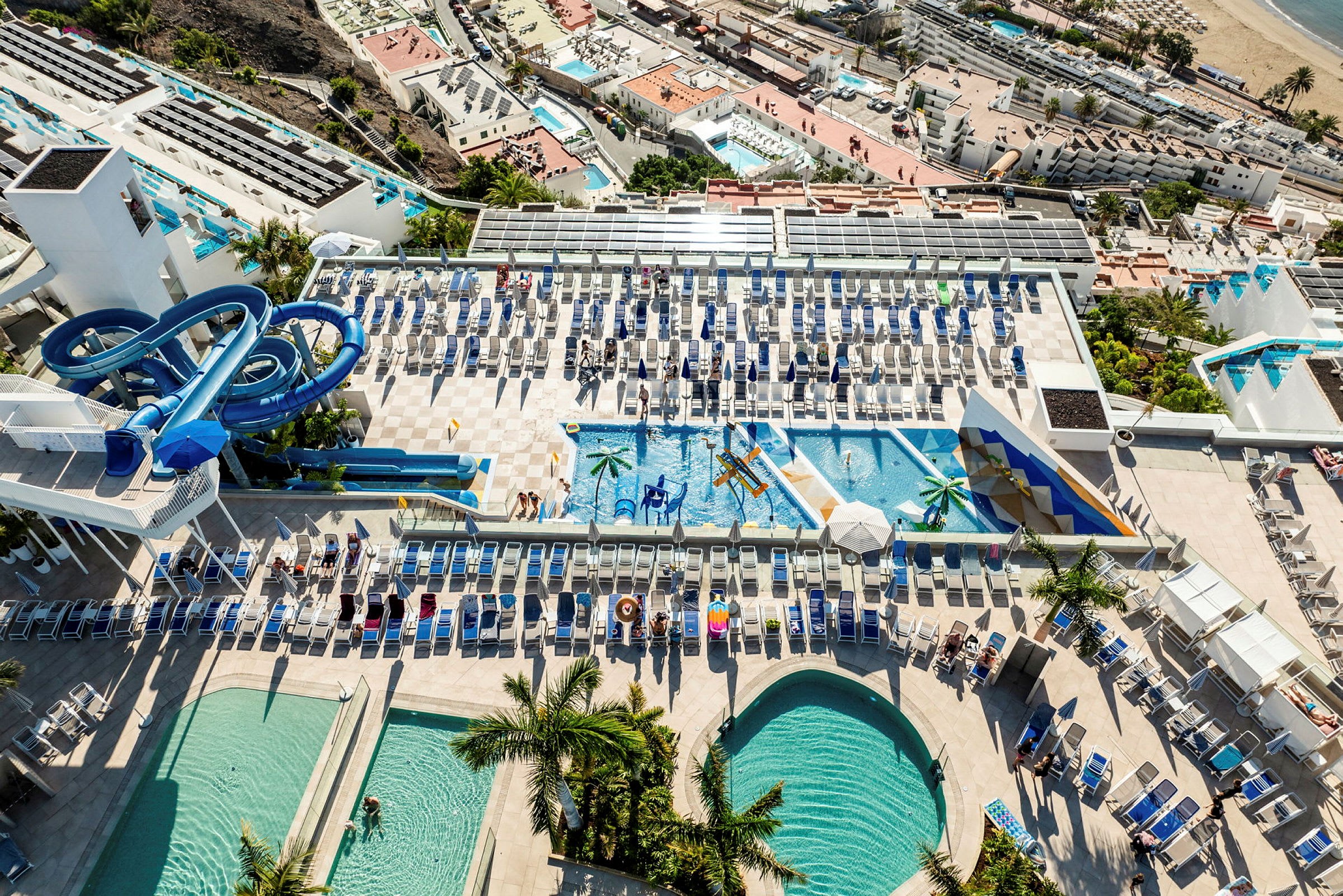 Flygbild av en resort med flera pooler, solstolar och vattenrutschbanor. (AI-genererad text)