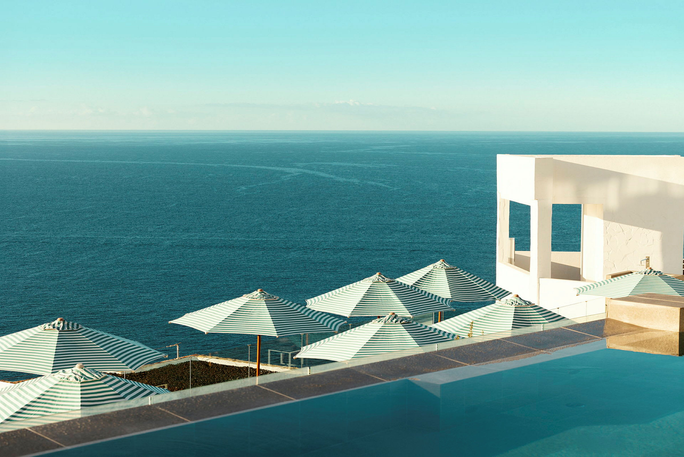 En bild med havet, grönstrimmiga parasoller och en vit byggnad vid en pool. (AI-genererad text)