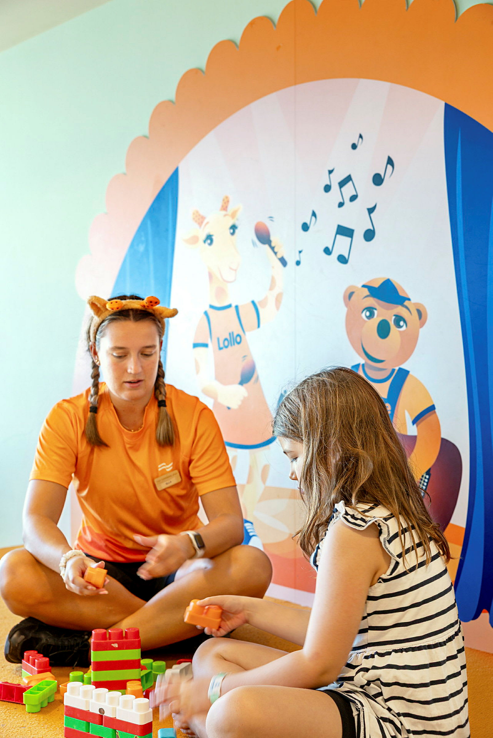Ett rum med barnleksaker där två personer leker med byggklossar, nedanför en färgglad vägg med illustrationer av barnfigurer. (AI-genererad text)