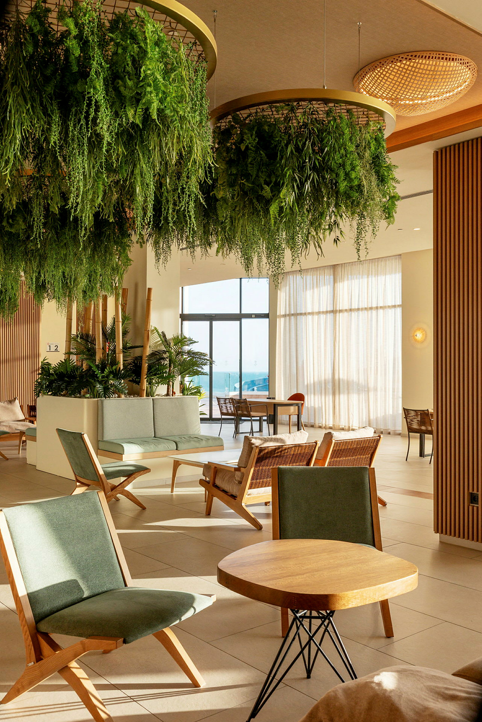 En hotellobby med gröna växter hängande från taket, sittgrupper med trästolar och bord, och glasdörrar som leder ut till en solig terrass. (AI-genererad text)