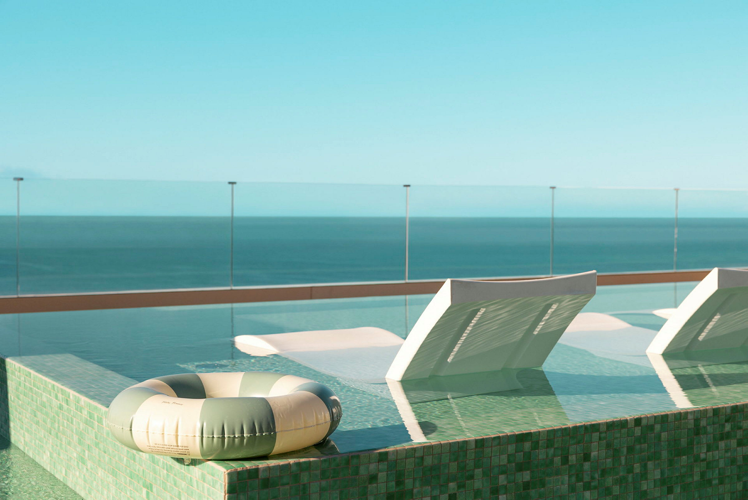 Utsikt över en pool med solstolar och badring vid ett glasräcke mot havet. (AI-genererad text)