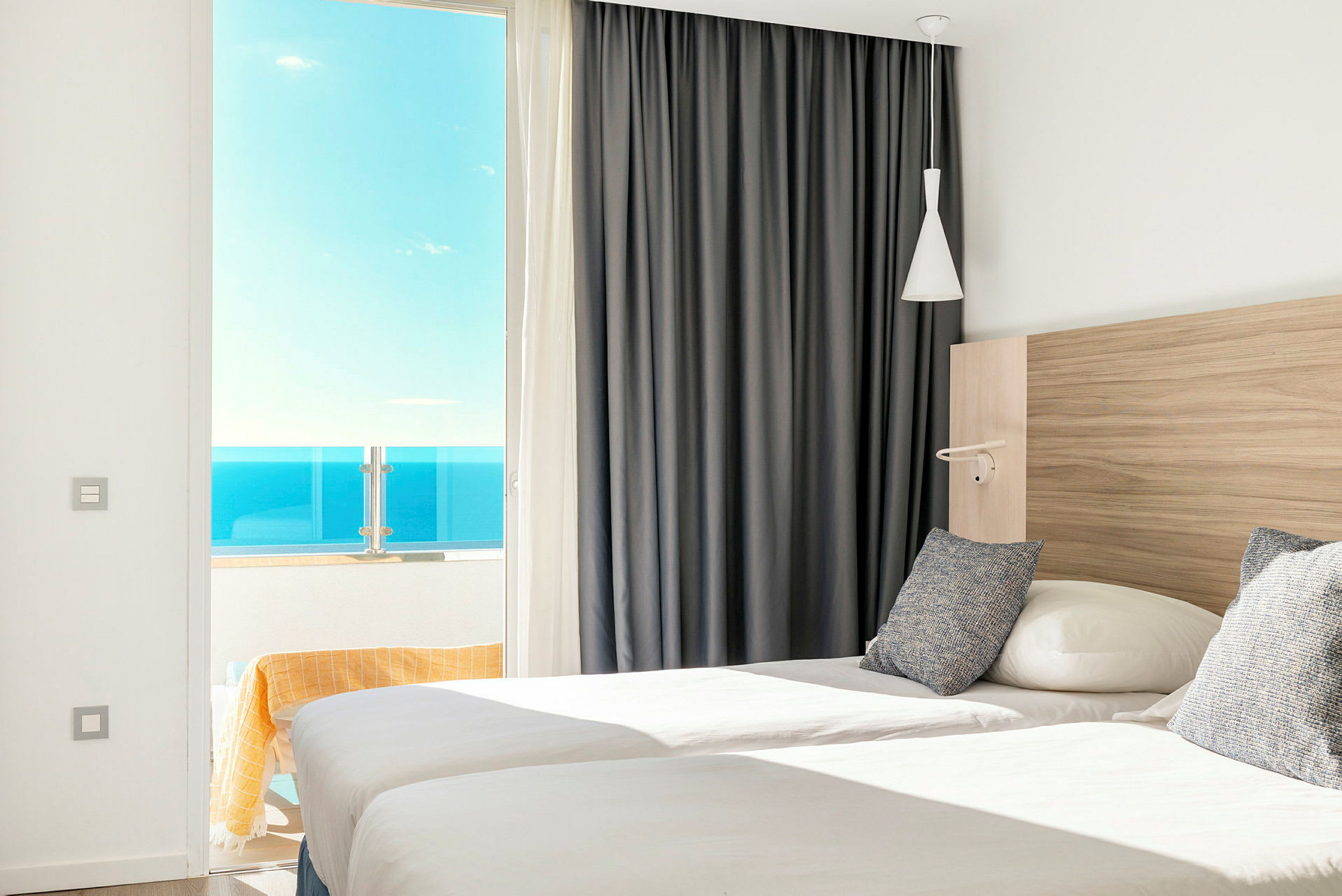 Ett hotellrum med två enkelsängar, öppet fönster mot en balkong med havsutsikt. (AI-genererad text)