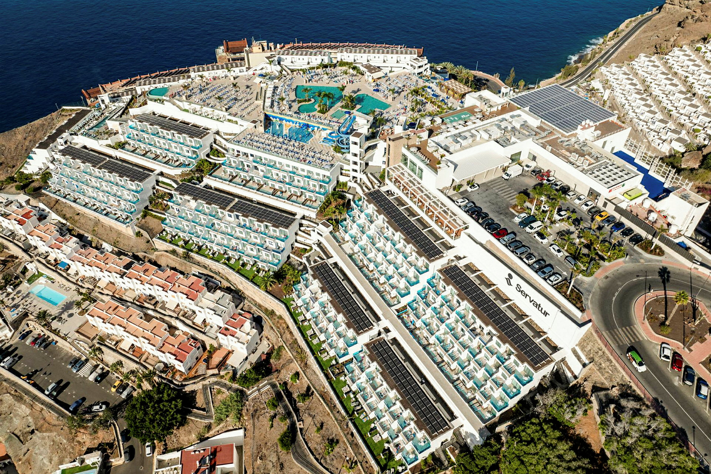 Flygfoto av ett stort hotellkomplex vid havet med poolområde och omgivande bostadshus. (AI-genererad text)
