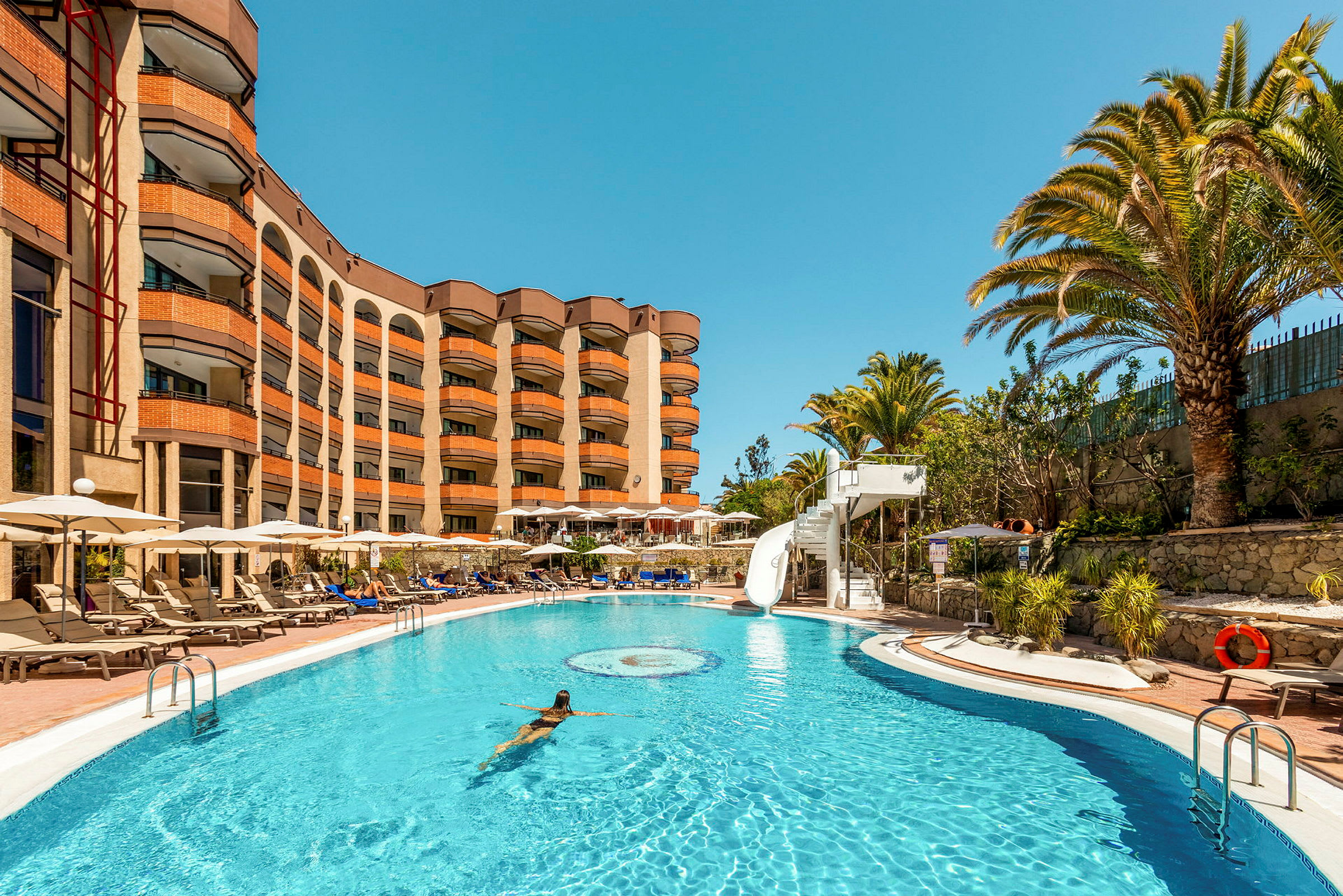 En pool med en kvinna som simmar, omgiven av solstolar och ett hotell i bakgrunden. (AI-genererad text)