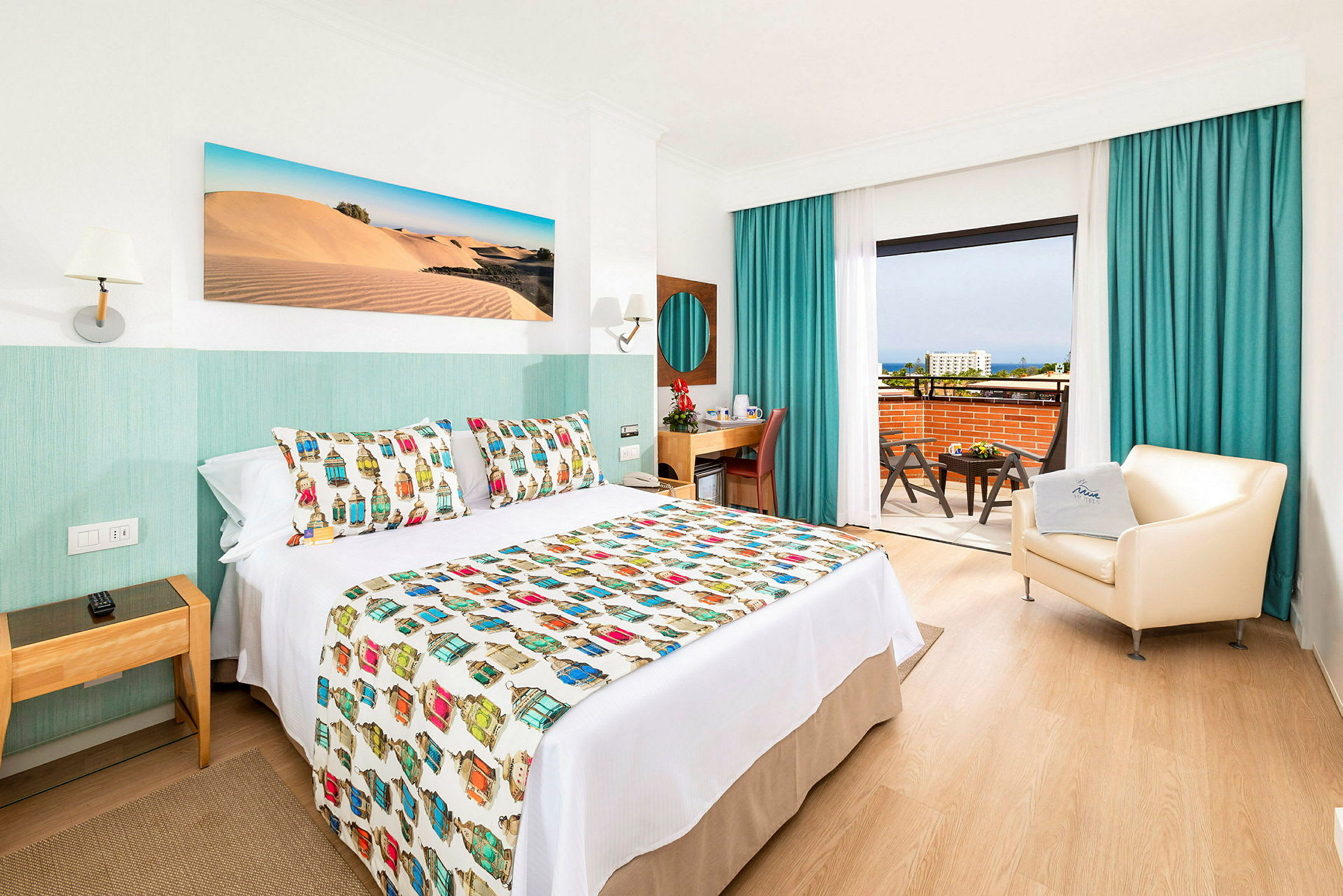 Hotelrum med dekorativt överkast, fåtölj och skrivbord samt utsikt från balkongen. (AI-genererad text)