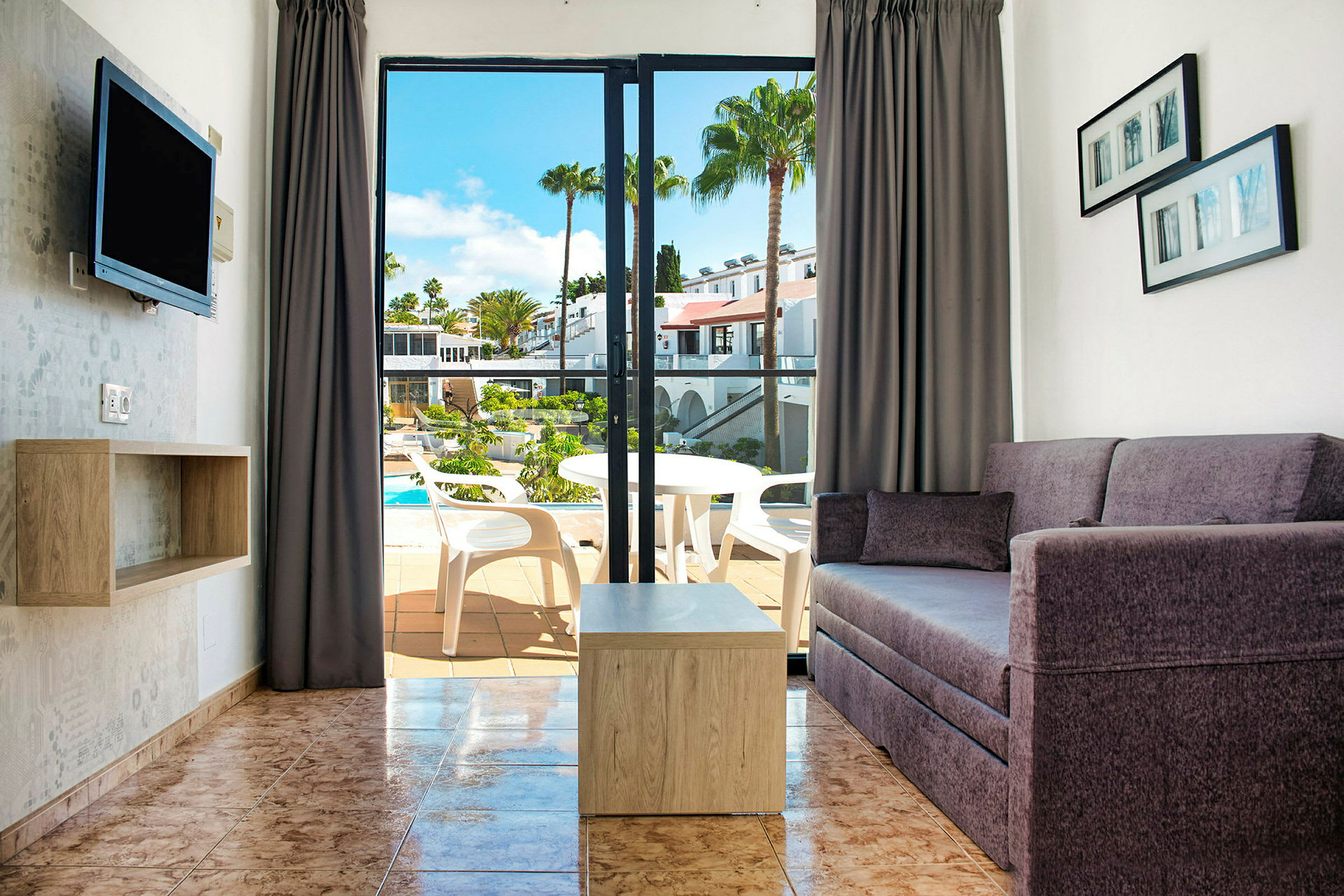 Vy av ett vardagsrum med soffa och bord samt en öppning till balkong med utsikt över en pool. (AI-genererad text)