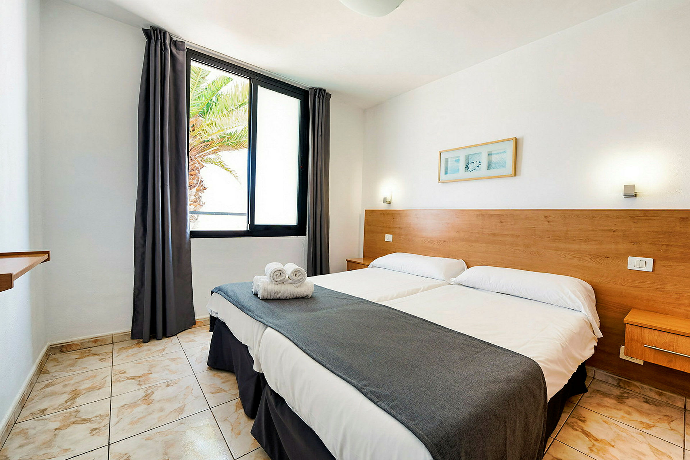 Hotellrum med två enkelsängar, sängarna är bäddade med vita lakan och ihoprullade handdukar ligger på ena sängen. Ett fönster med mörka gardiner visar en palm utanför. (AI-genererad text)