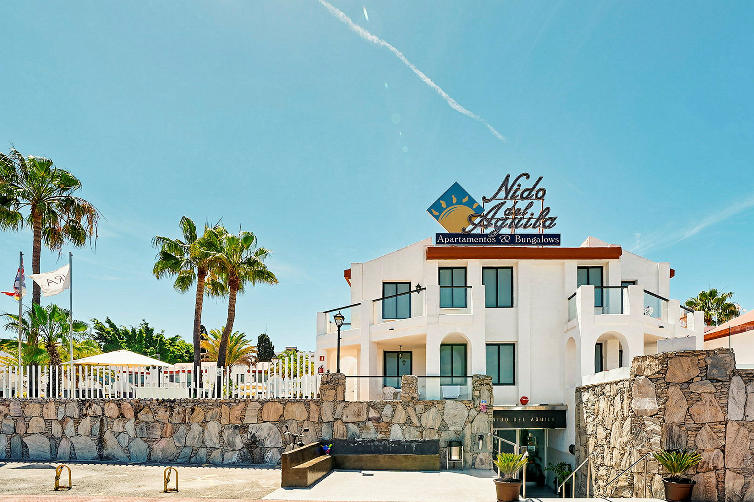 Ett semesterhotell med vita väggar och en skylt som lyder 'Nido del Águila', omgiven av palmer. (AI-genererad text)