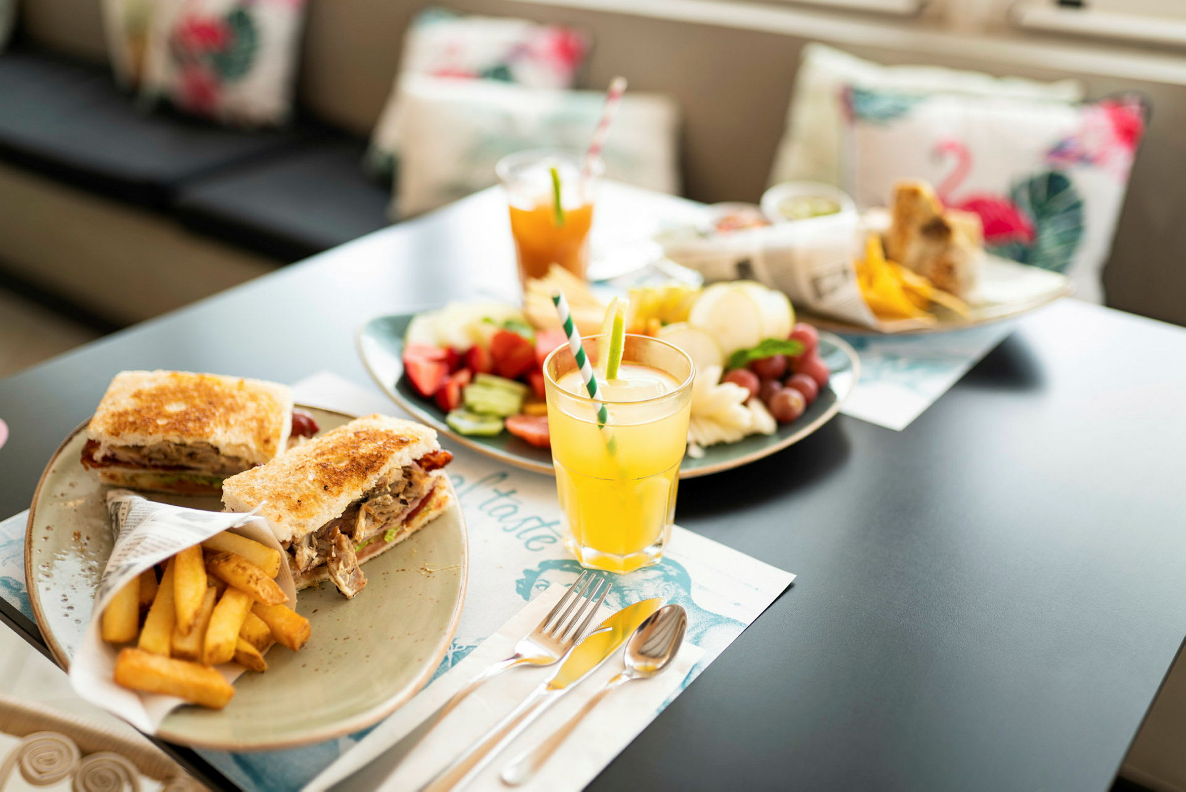 En bild på ett bord med olika tallrikar innehållande mat och dryck, inklusive smörgåsar, pommes frites, frukt och juice. (AI-genererad text)