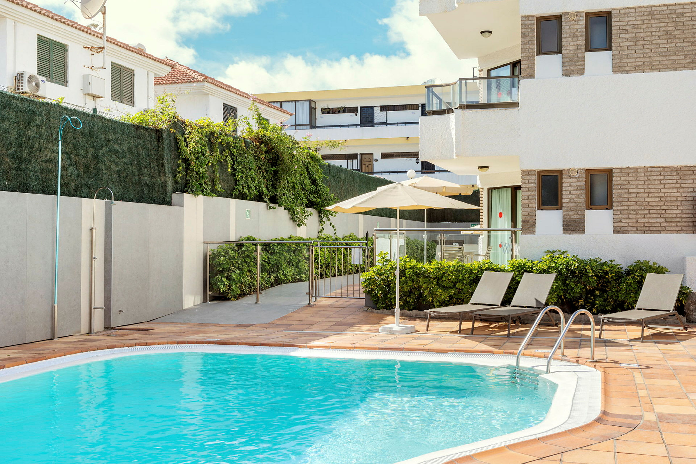 Bild av ett litet poolområde med solstolar och en parasoll vid en turkosblå pool, omgiven av vegetation och byggnader. (AI-genererad text)