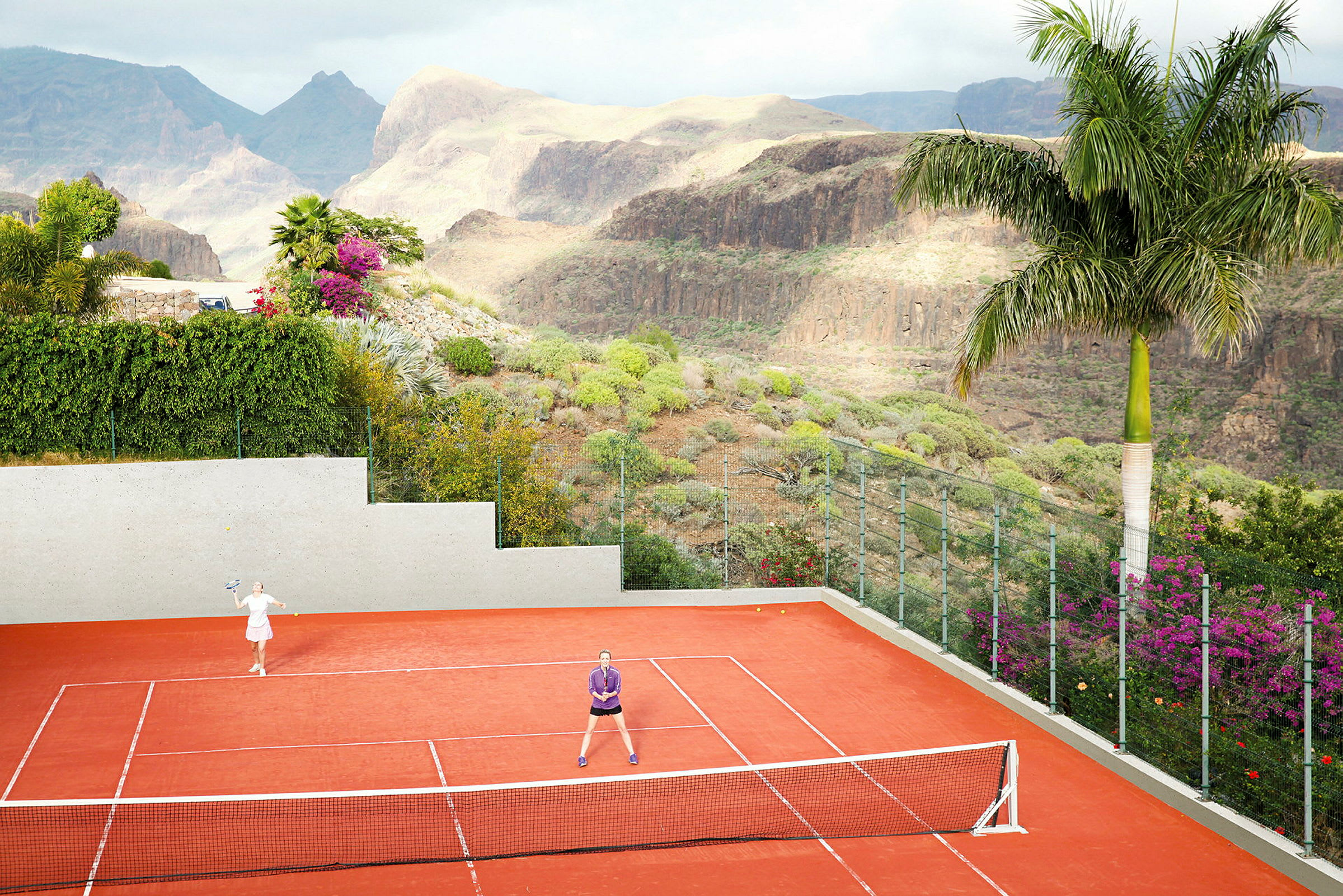 Två personer spelar tennis på en röd tennisplan, omgivna av en grön dal. (AI-genererad text)