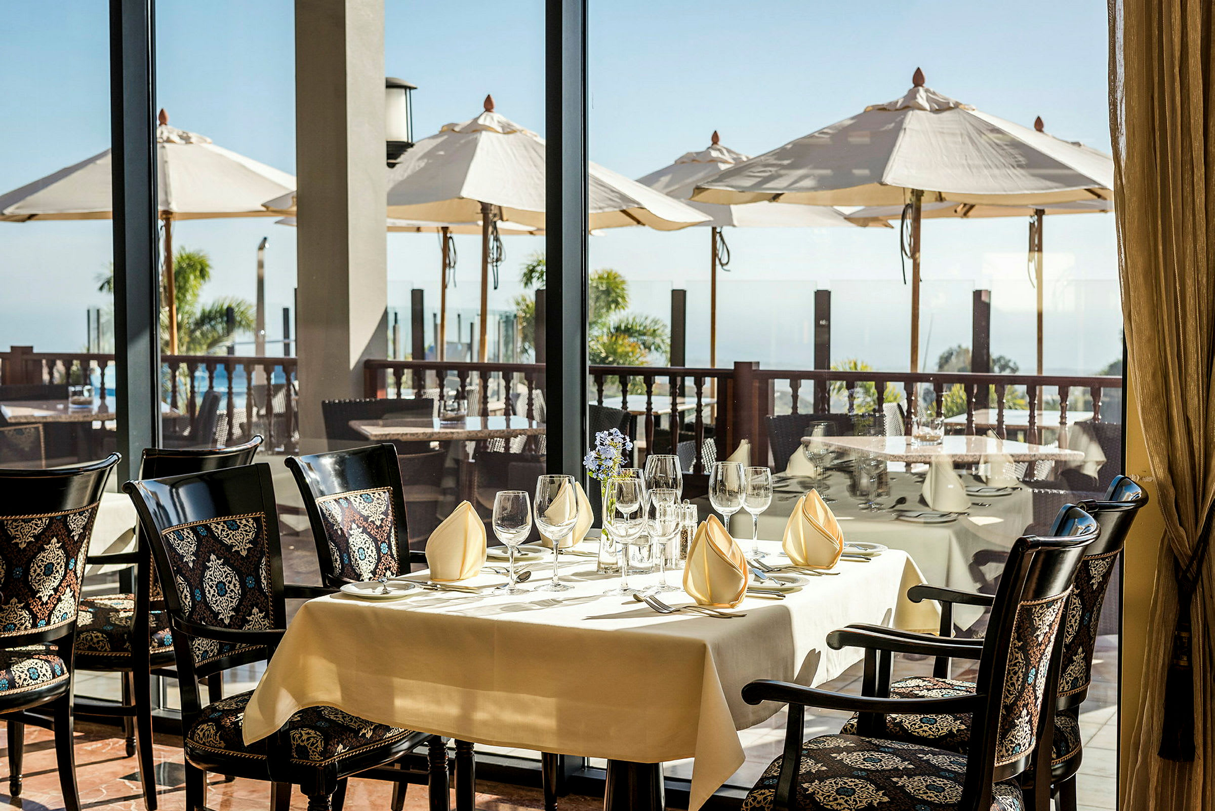 Ett dukat bord i en restaurang med utsikt över en terrass med parasoller och havet. (AI-genererad text)