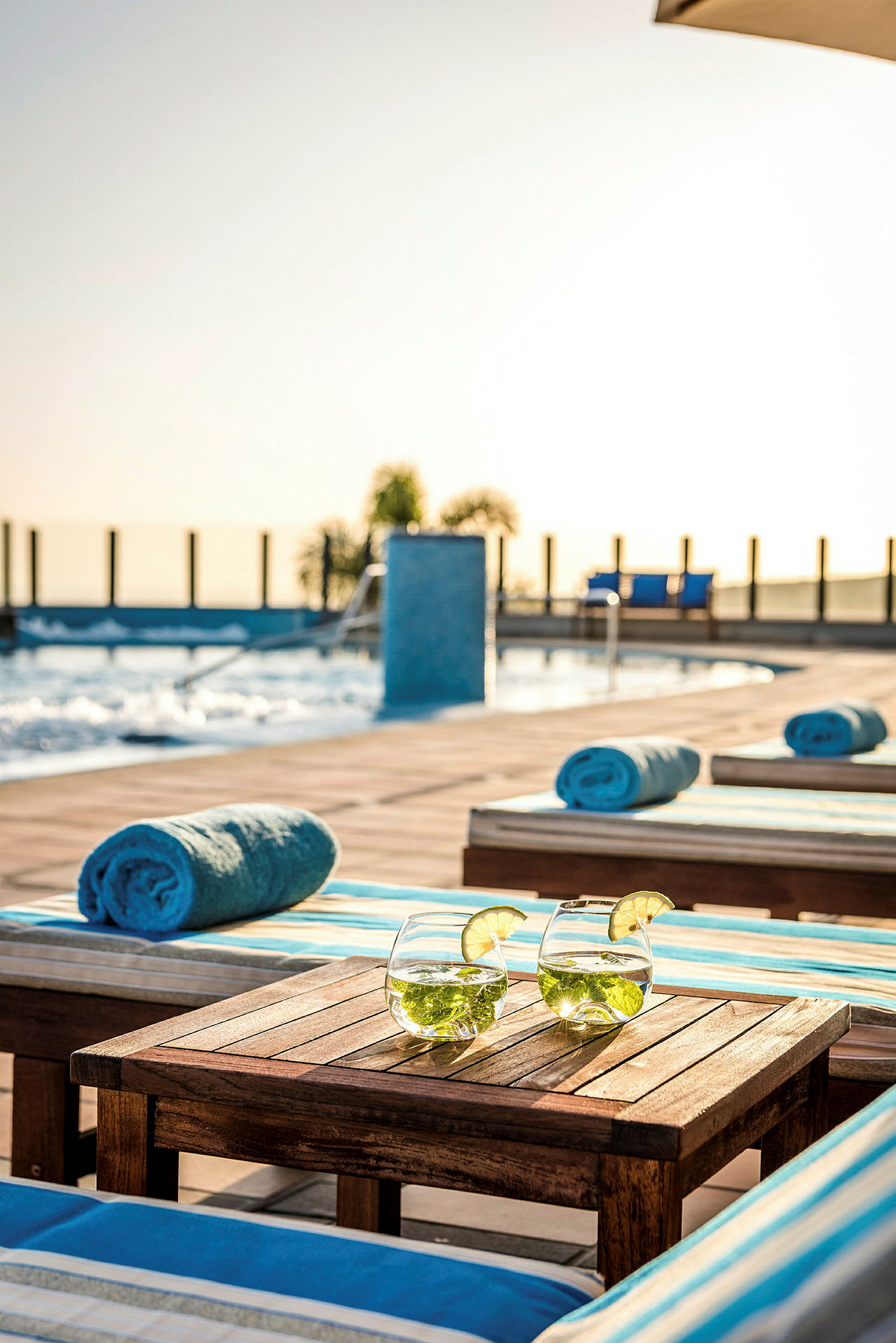 Två glas med dryck, dekorerade med lime, står på ett bord vid solstolar och handdukar nära en pool. (AI-genererad text)