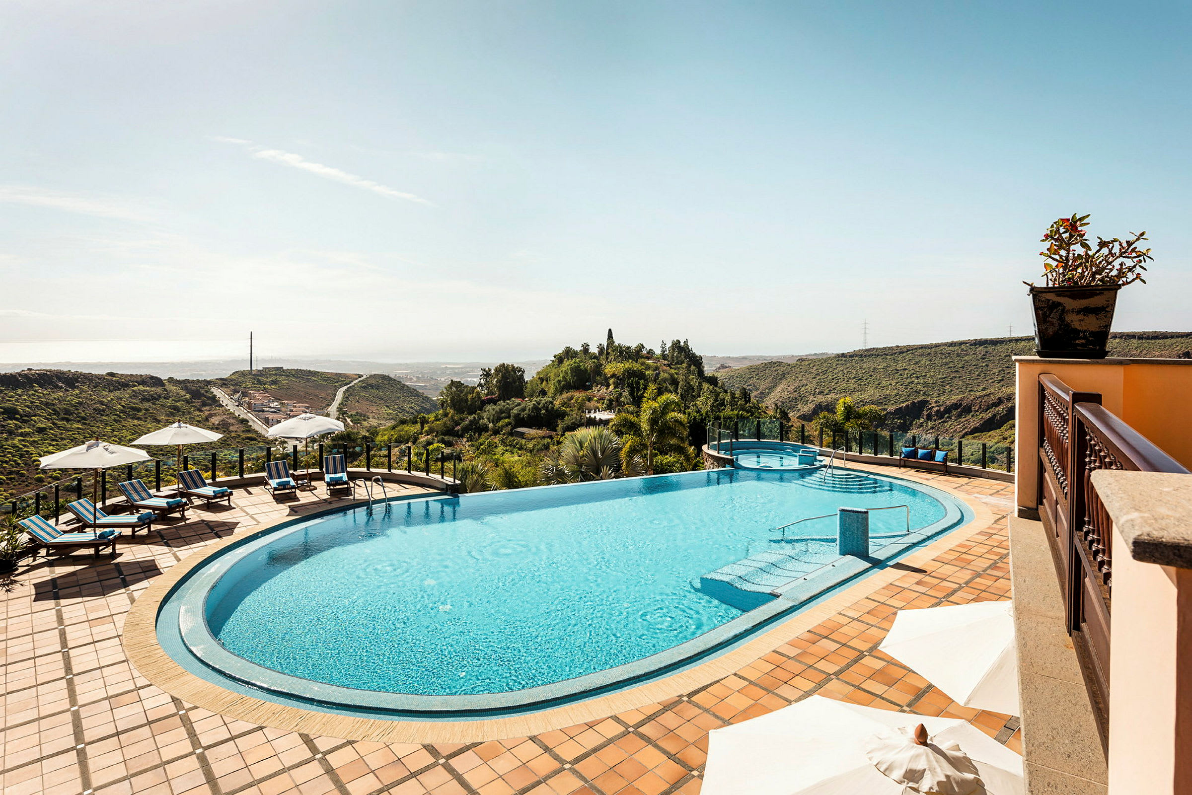 Utsikt över en infinitypool med solstolar och parasoller på en terrass, omgiven av grönskande landskap. (AI-genererad text)