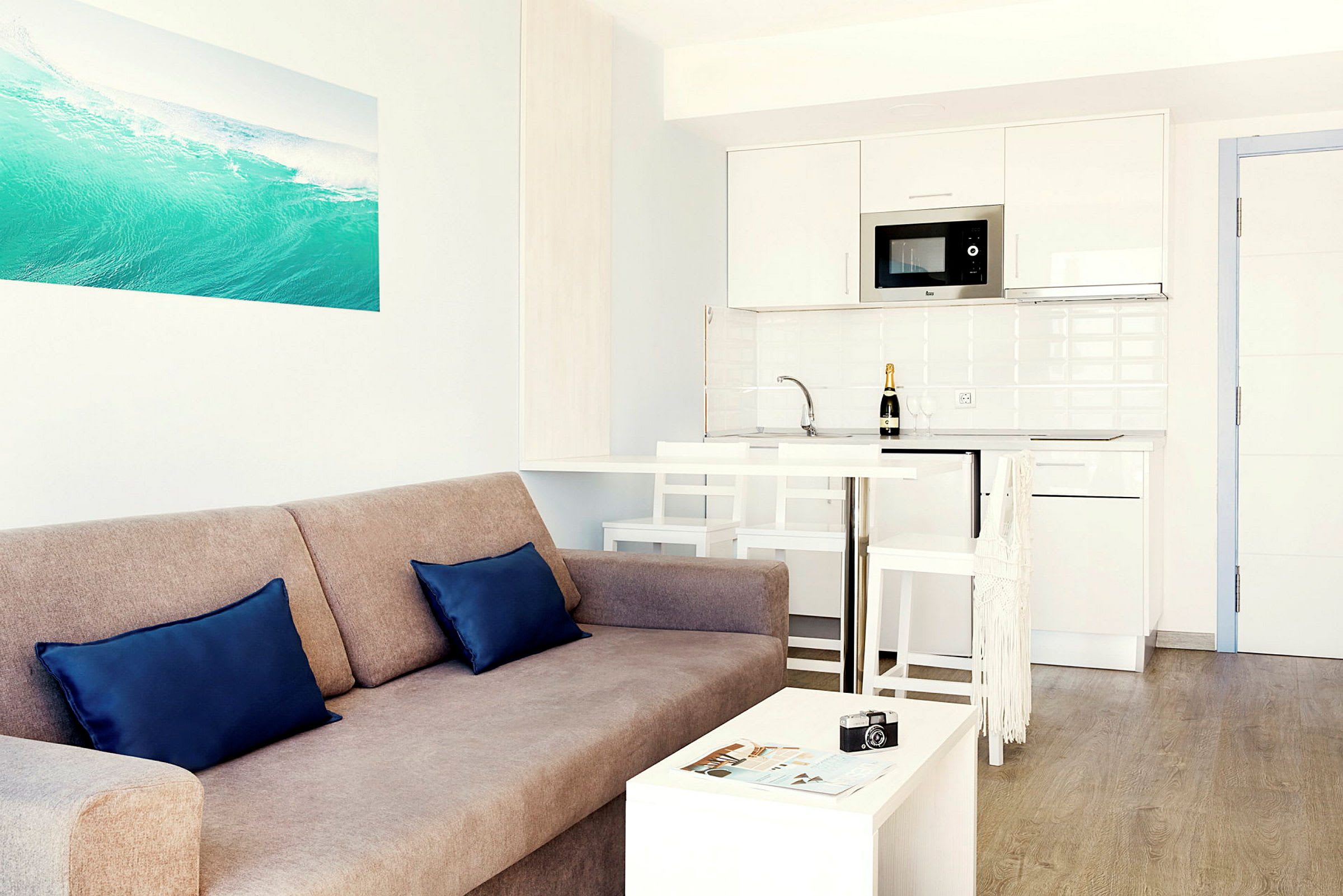 Ett ljust och modernt vardagsrum med en soffa, två kuddar, ett litet bord, samt ett kök med vit inredning i bakgrunden. (AI-genererad text)
