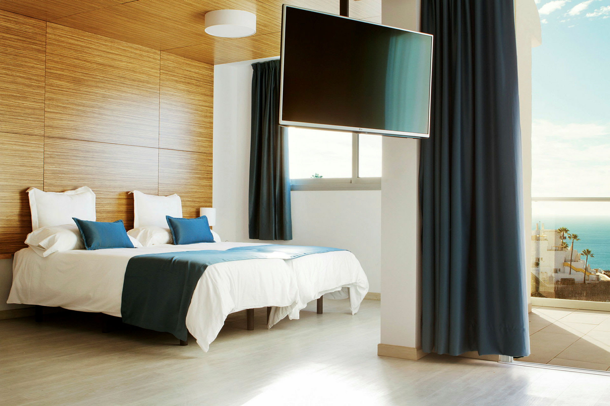 Ett hotellrum med två bäddar, blå sängkläder, en väggmonterad TV och balkong med havsutsikt. (AI-genererad text)