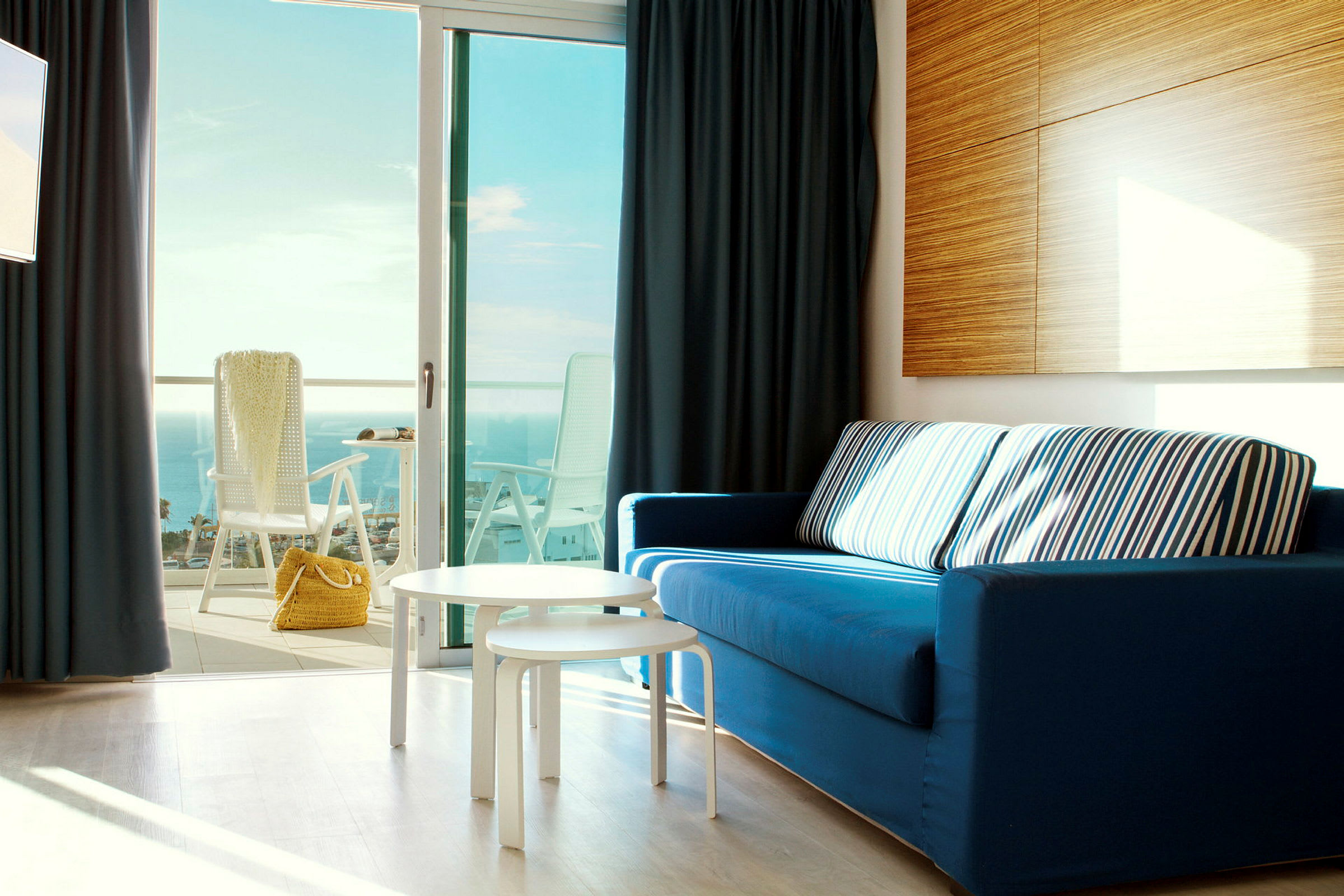 En interiörbild med en blå soffa, vita bord och en öppen balkongdörr med utsikt över havet. (AI-genererad text)