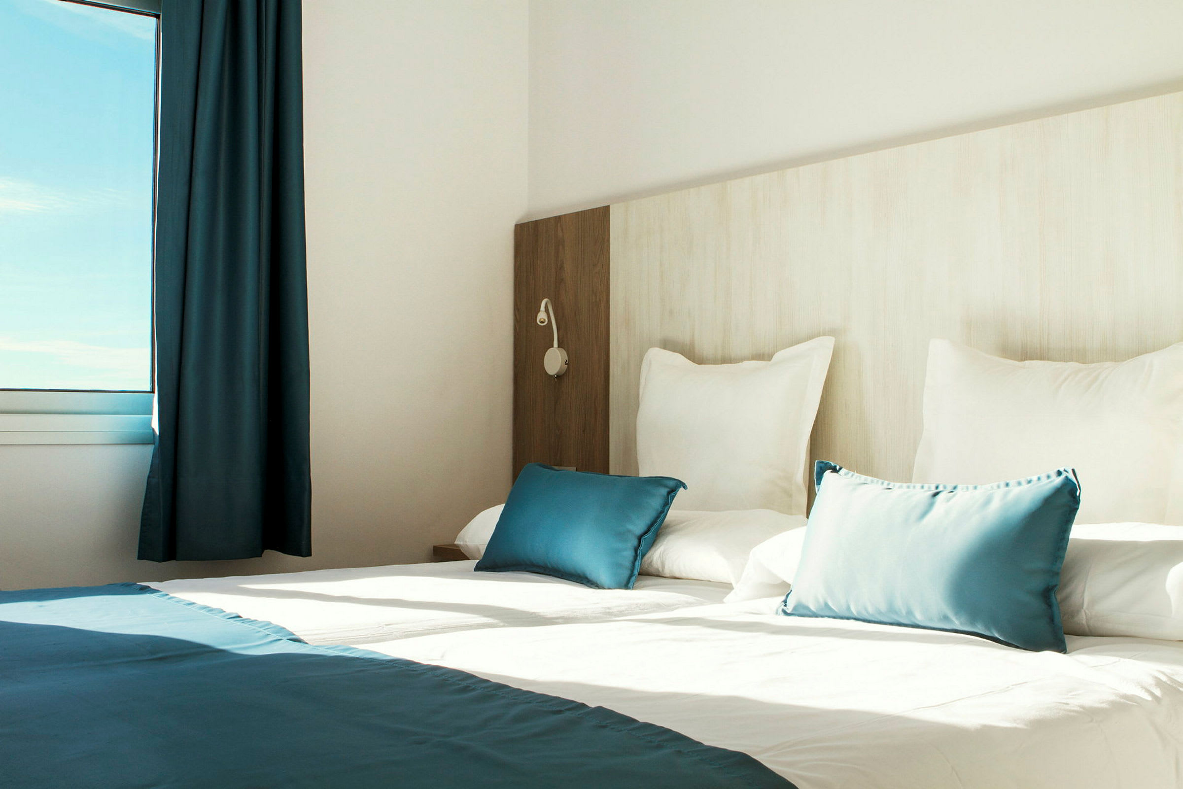Interiör av ett ljus hotellrum med en uppbäddad säng, vita och blå kuddar samt ett fönster som släpper in naturligt ljus. (AI-genererad text)