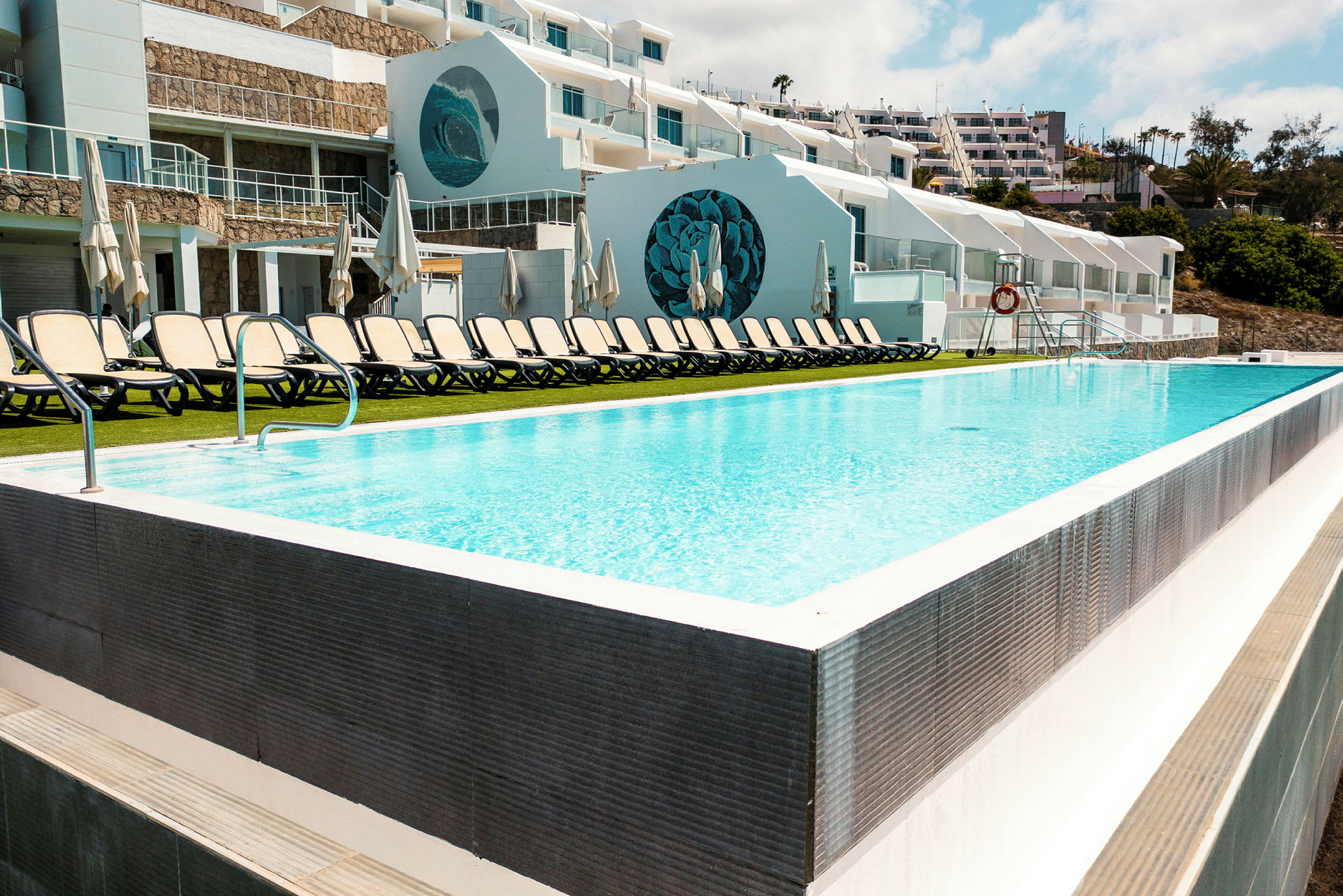 En swimmingpool med solsängar och vit byggnad i bakgrunden. (AI-genererad text)