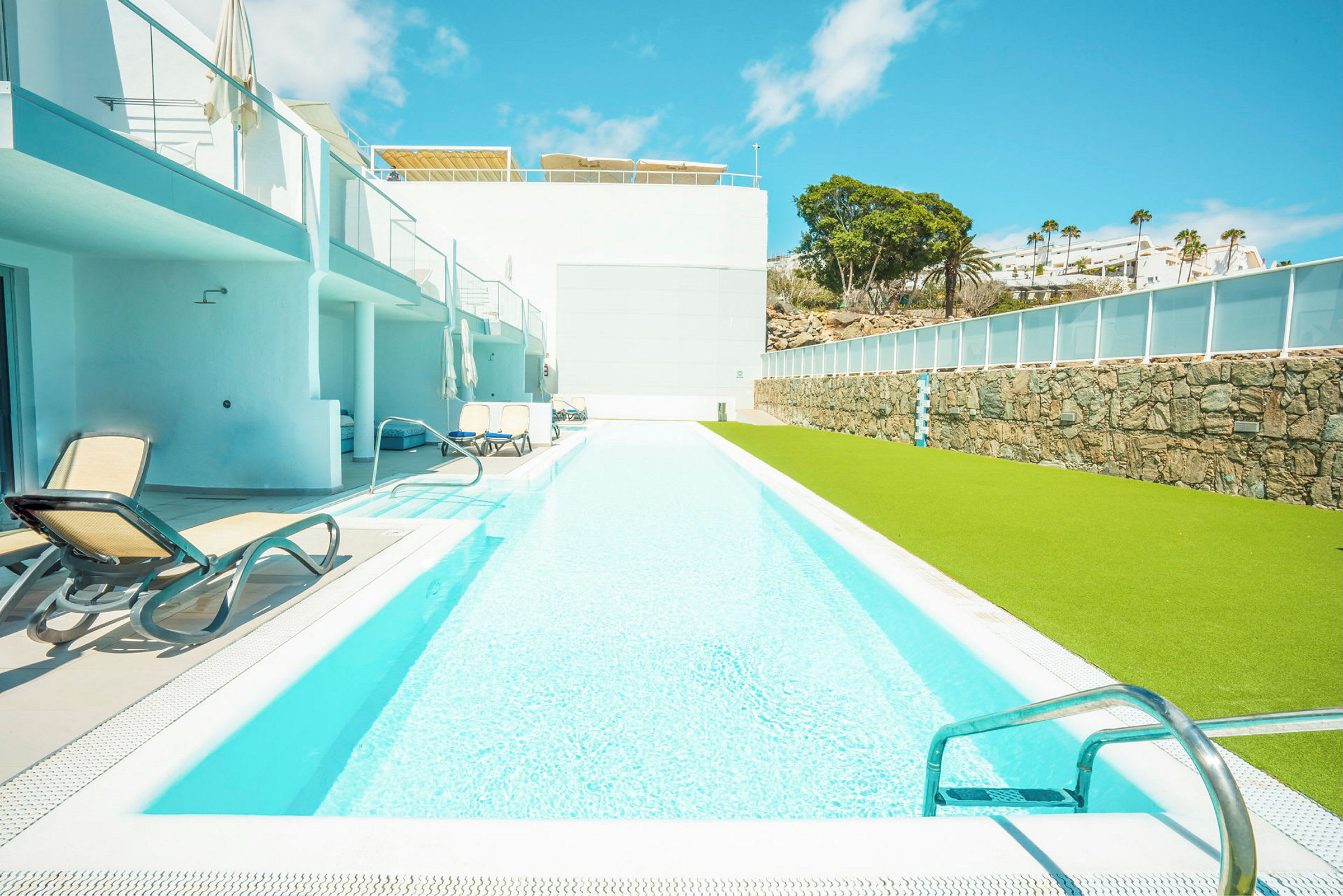 En lång swimmingpool med solstolar vid sidan och en grön gräsyta. (AI-genererad text)