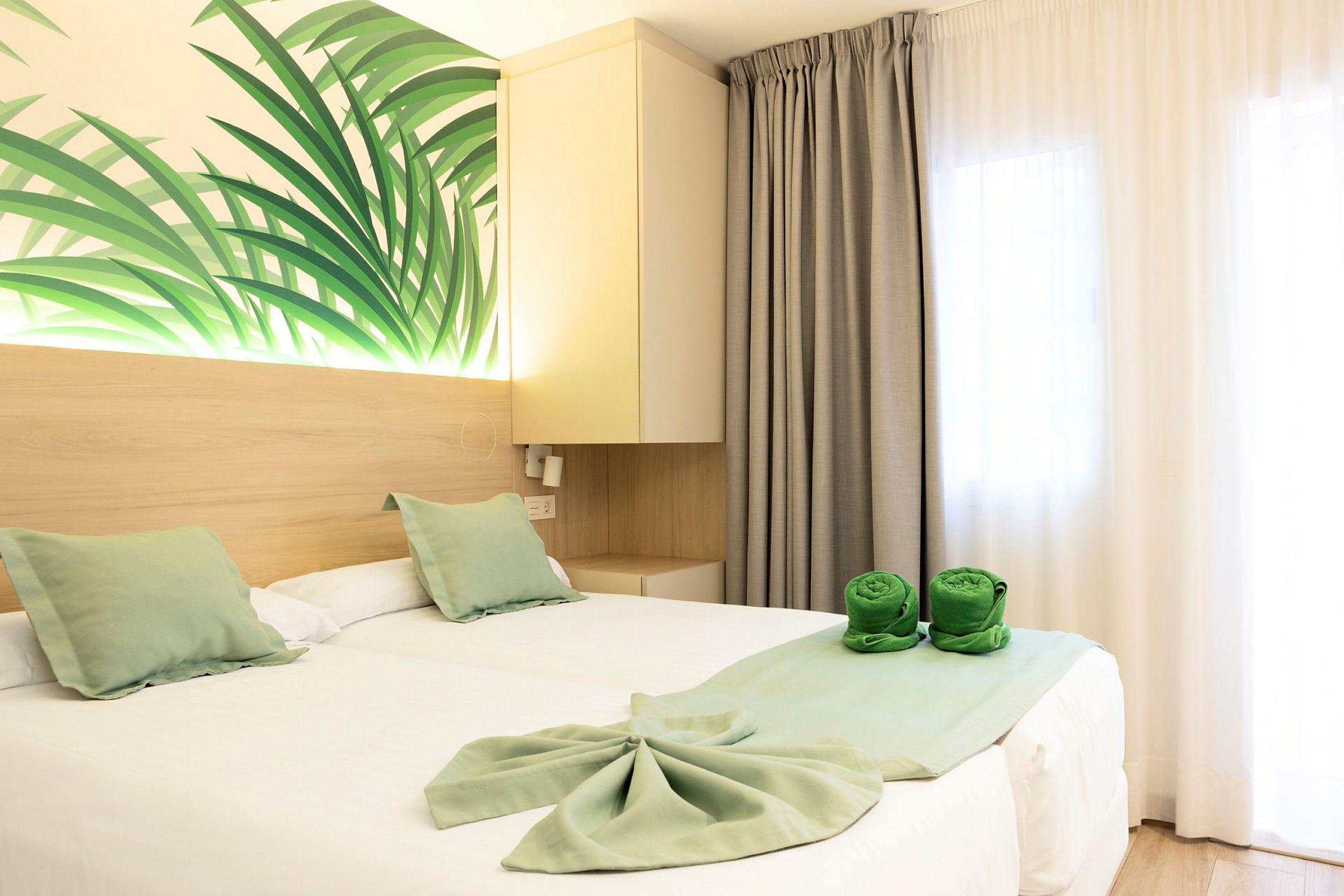 Ett hotellrum med två enkelsängar täckta av vita lakan och gröna dekorativa kuddar. Väggdekoration med bladmotiv samt gröna hoprullade handdukar på sängen. (AI-genererad text)