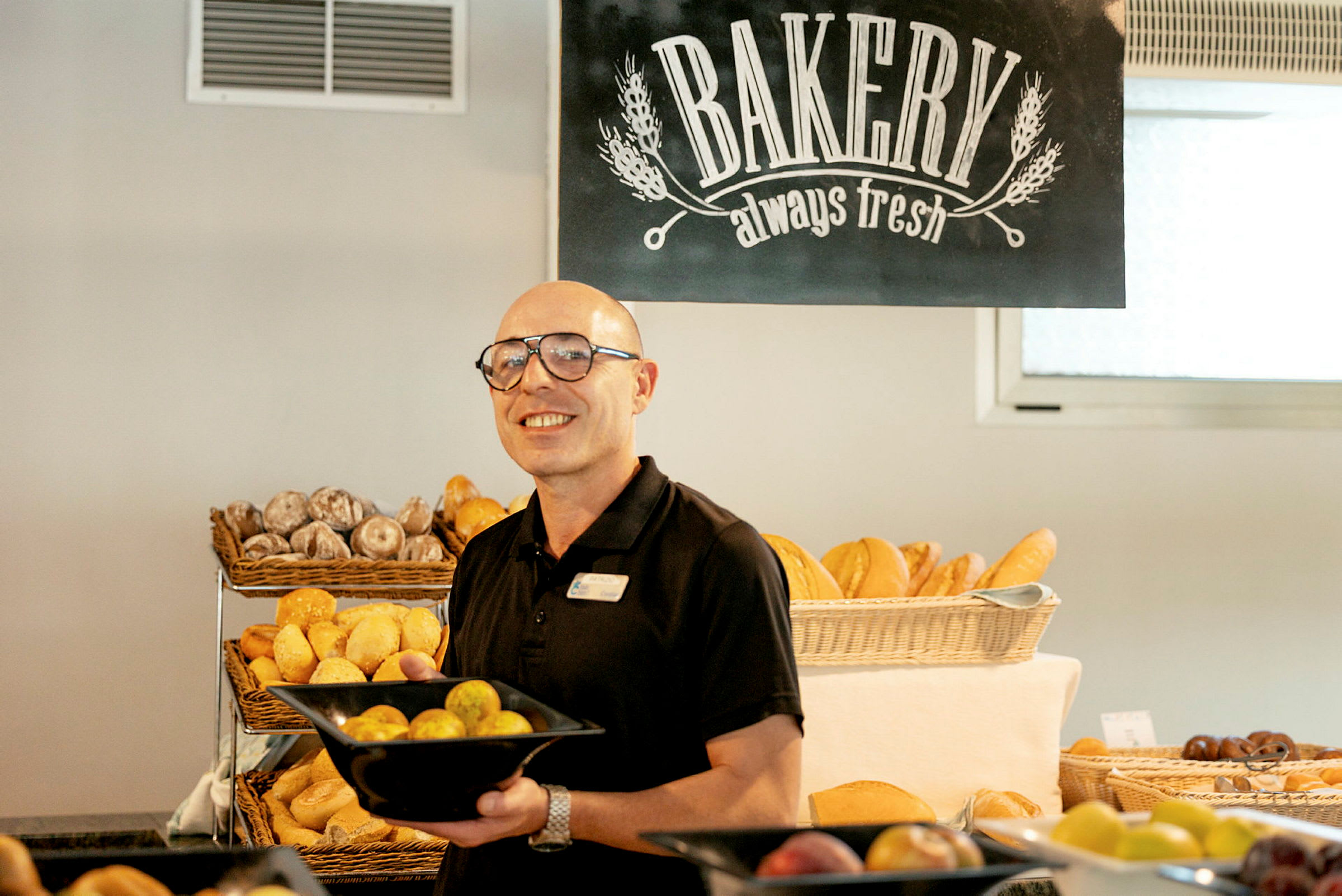 En person håller en svart skål fylld med bröd i ett bageri, med andra bakverk och ett skylt med texten 'Bakery always fresh' synliga i bakgrunden. (AI-genererad text)