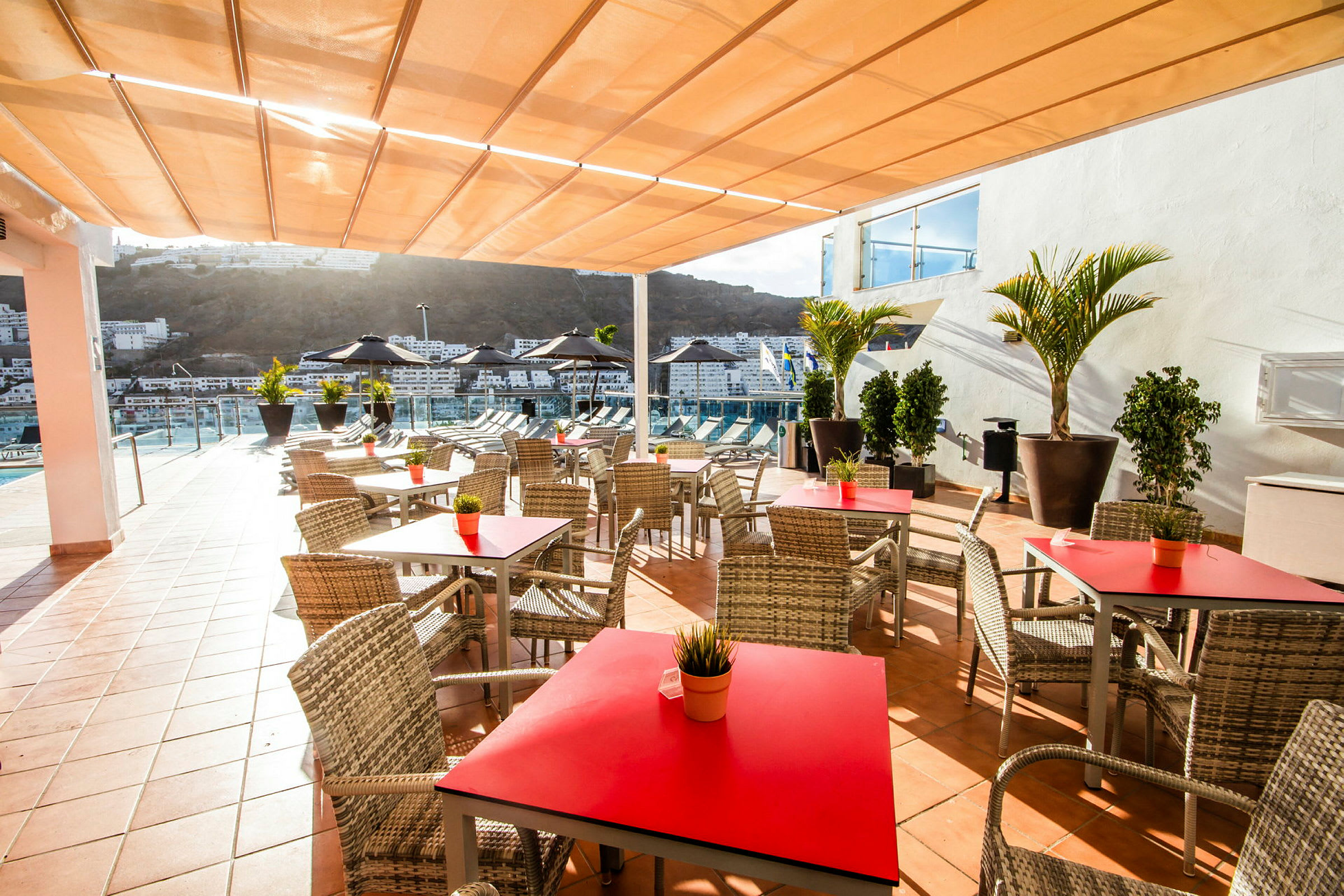 Terrass med bord och stolar, utsikt över pool och byggnader i bakgrunden. (AI-genererad text)
