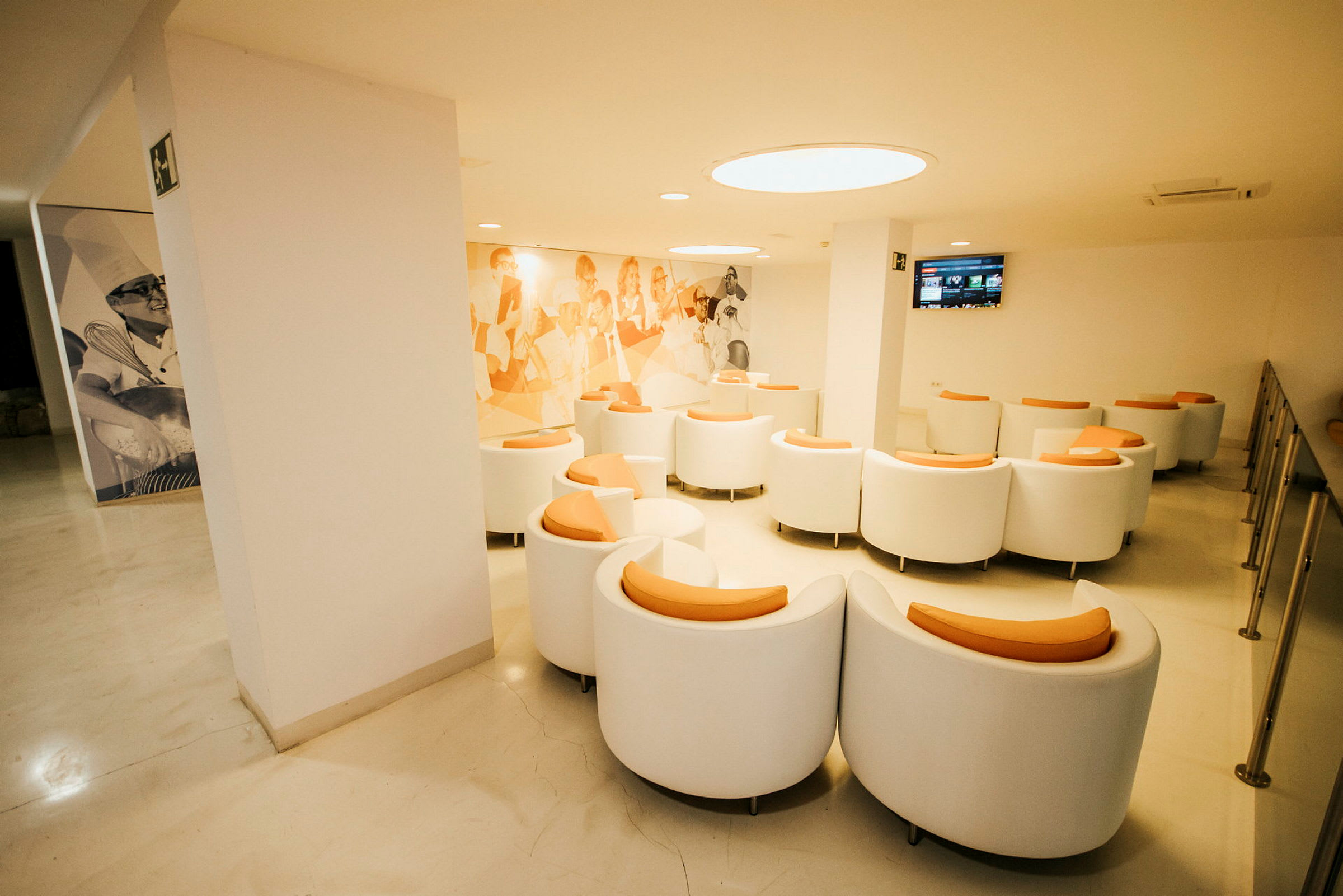 Ett väntområde med vita stolar med orange sittdynor, väggprydnader och en väggmonterad skärm. (AI-genererad text)
