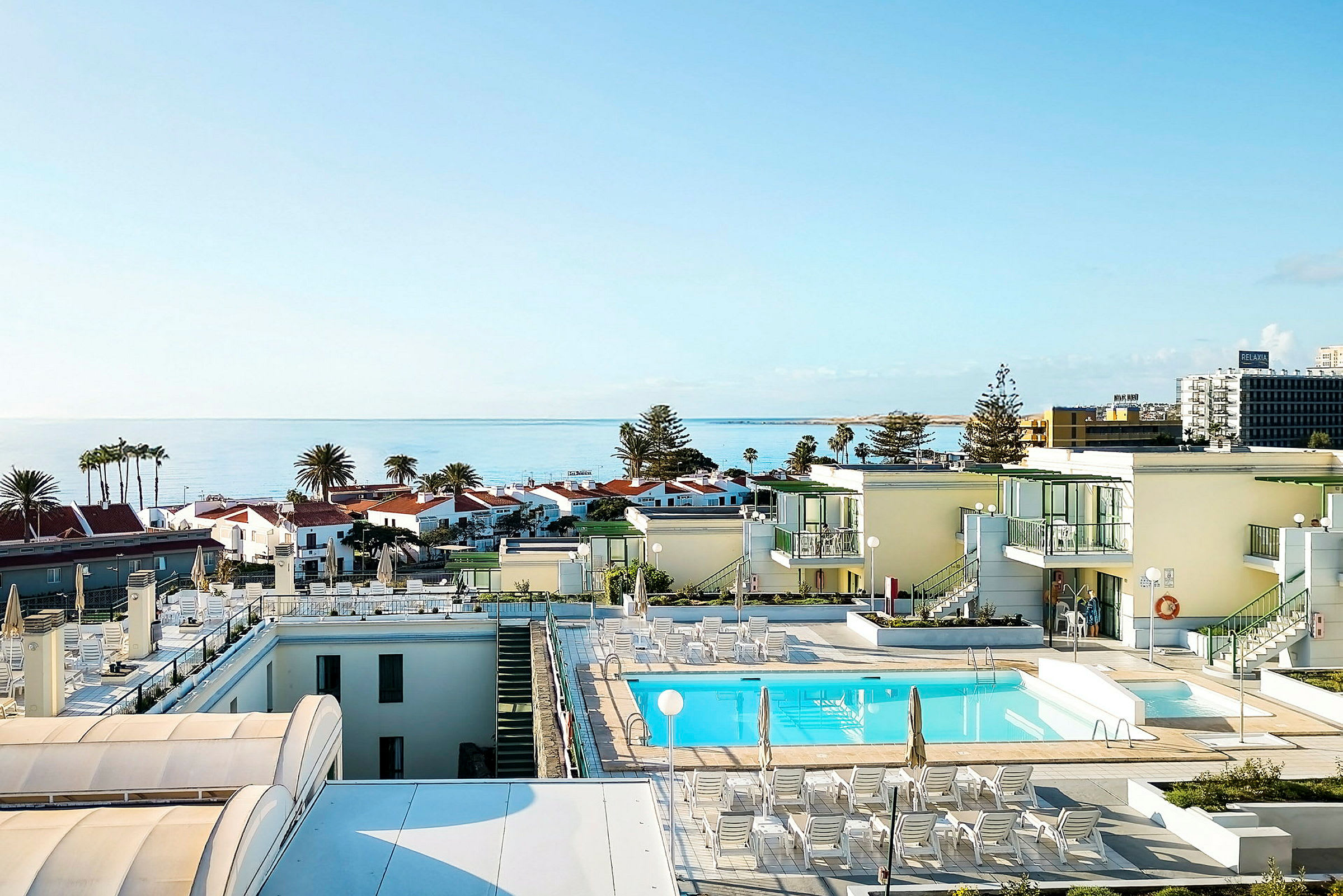 Vy över en pool på ett hotell med havet och byggnader i bakgrunden. (AI-genererad text)