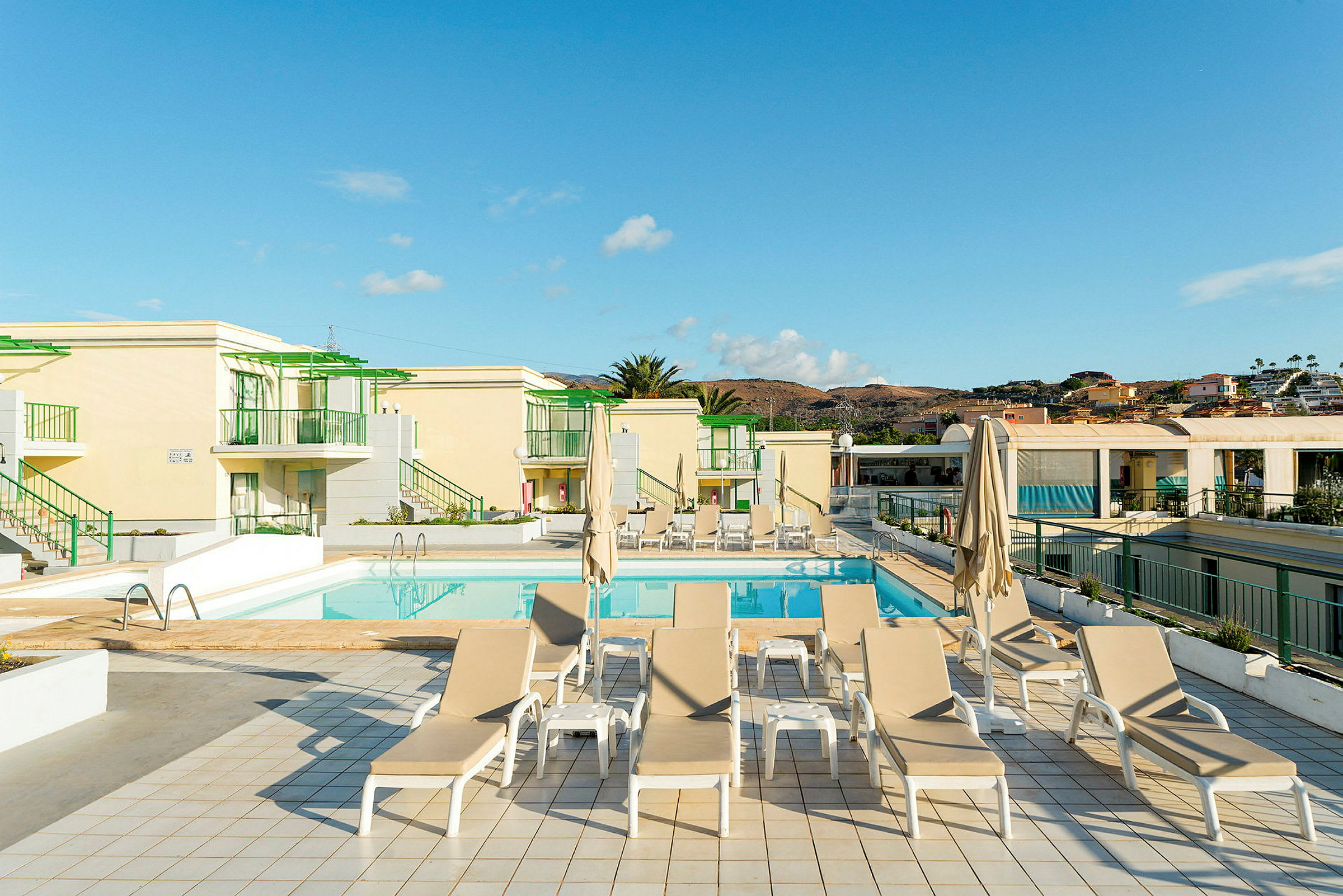 En solterrass vid en pool med solstolar och parasoller, omgiven av byggnader. (AI-genererad text)