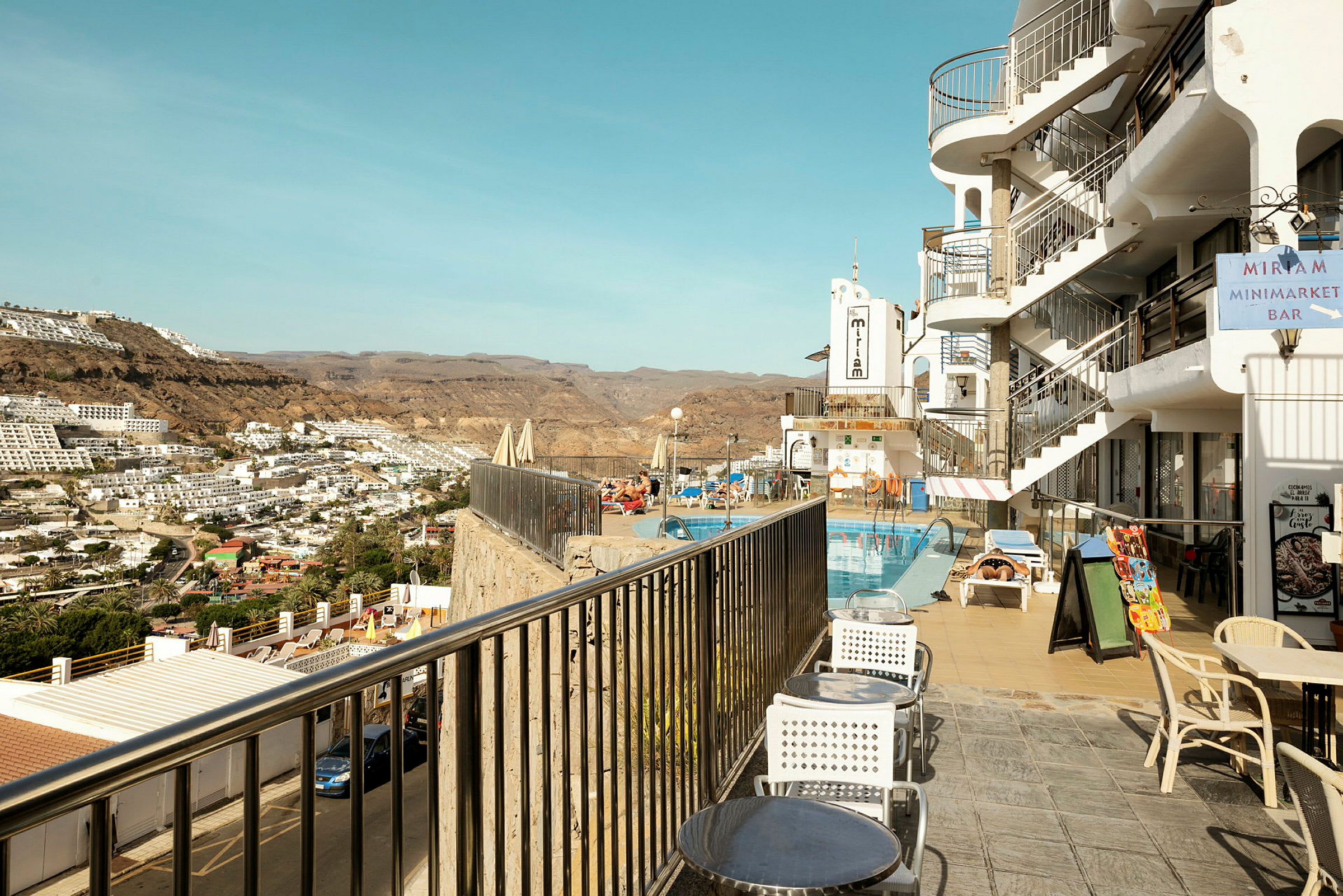 Bilden visar en bergsutsikt med vita byggnader i ett bostadsområde och ett hotell med pool och sittgrupp vid kanten. (AI-genererad text)