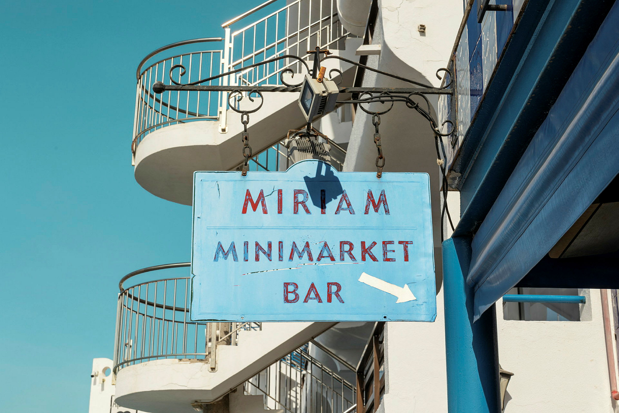 Skylt med texten 'Miriam Minimarket Bar' hängande på en byggnad. (AI-genererad text)
