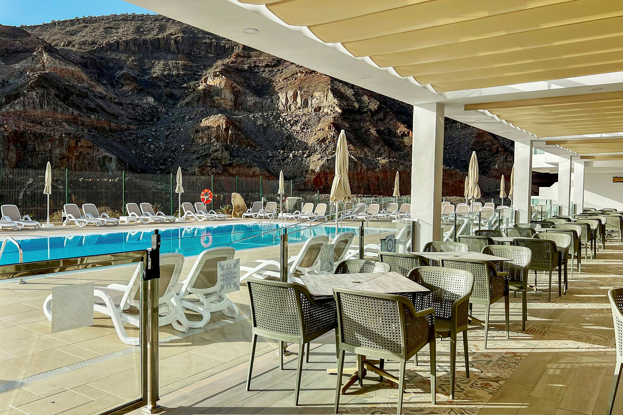 Utsikt över en pool med solstolar och parasoller framför en bergig bakgrund samt matplats med bord och stolar på en terrass. (AI-genererad text)