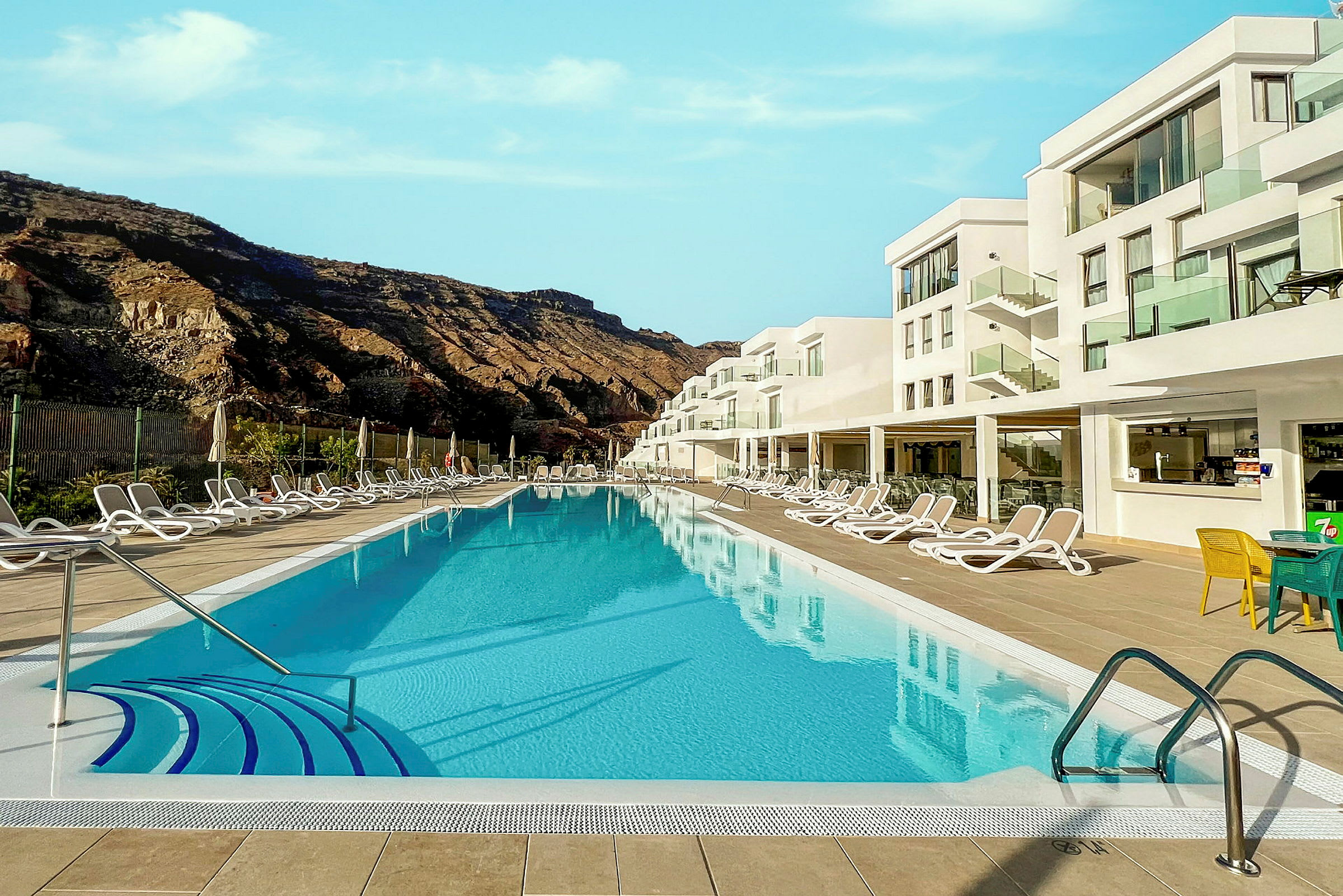 En pool omgiven av solstolar och en modern hotellbyggnad i bakgrunden. (AI-genererad text)