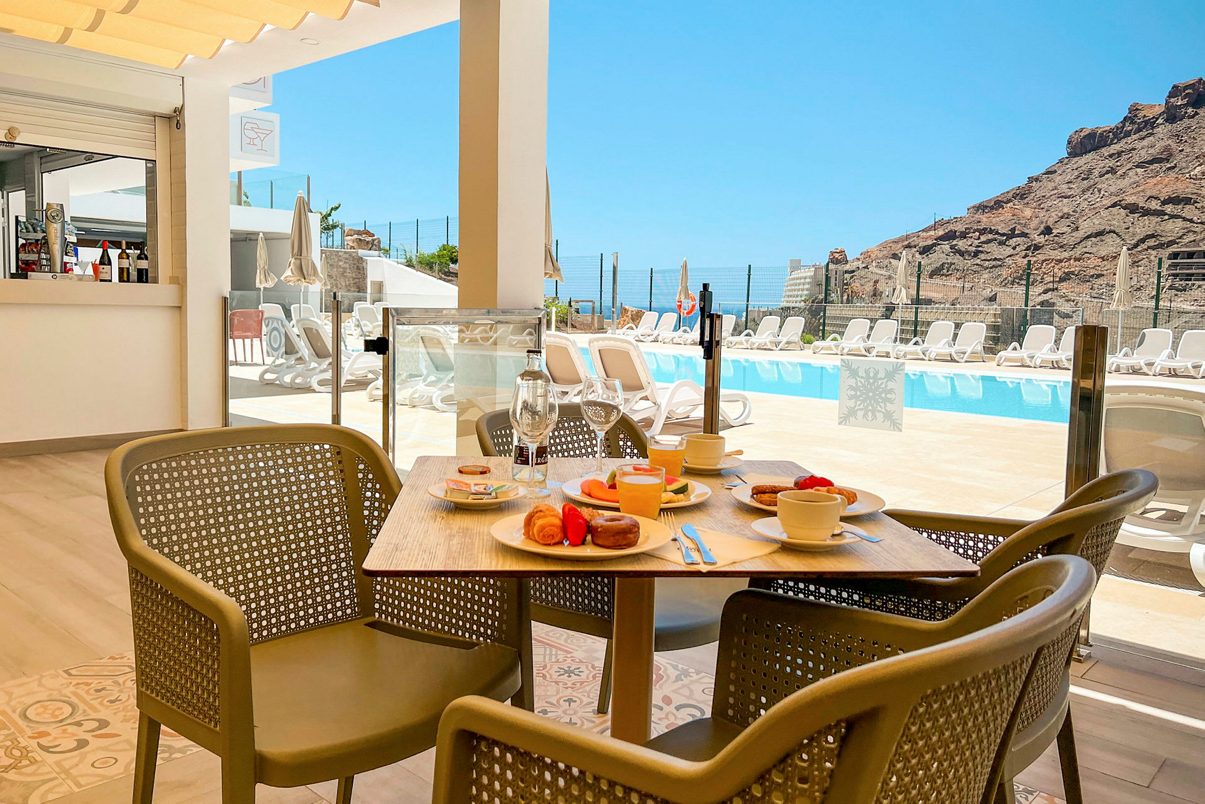 Ett bord med frukost och stolar framför en pool och berg i bakgrunden. (AI-genererad text)