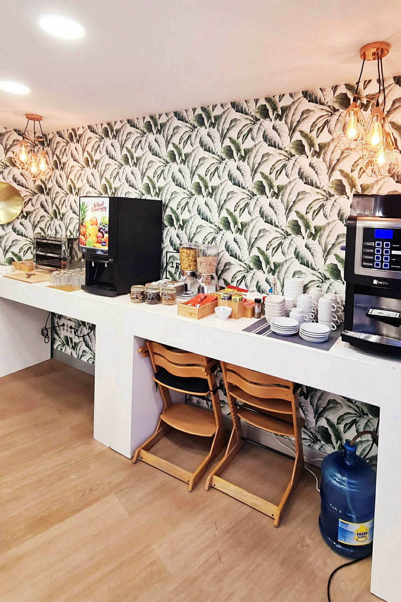 Bild av en frukostbuffé med kaffemaskin, juiceautomat, tallrikar, koppar och livsmedel presenterade på en vit köksbänk. Stol och vattenbehållare i förgrunden. (AI-genererad text)
