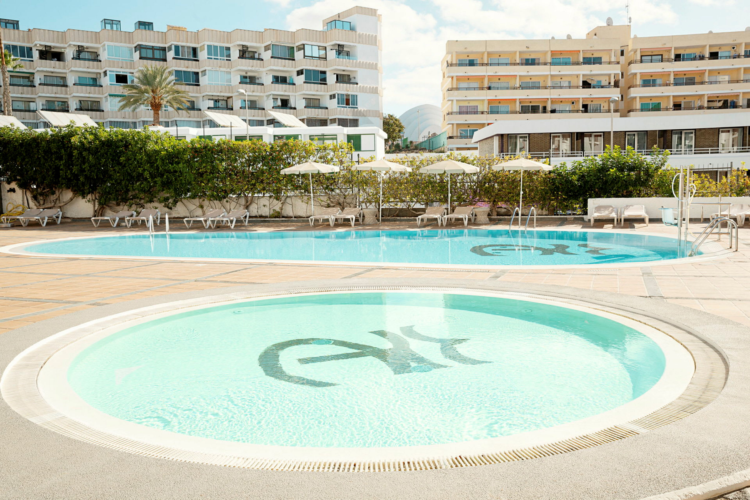 Två swimmingpooler omgivna av solstolar och parasoller, med hotellbyggnader i bakgrunden. (AI-genererad text)