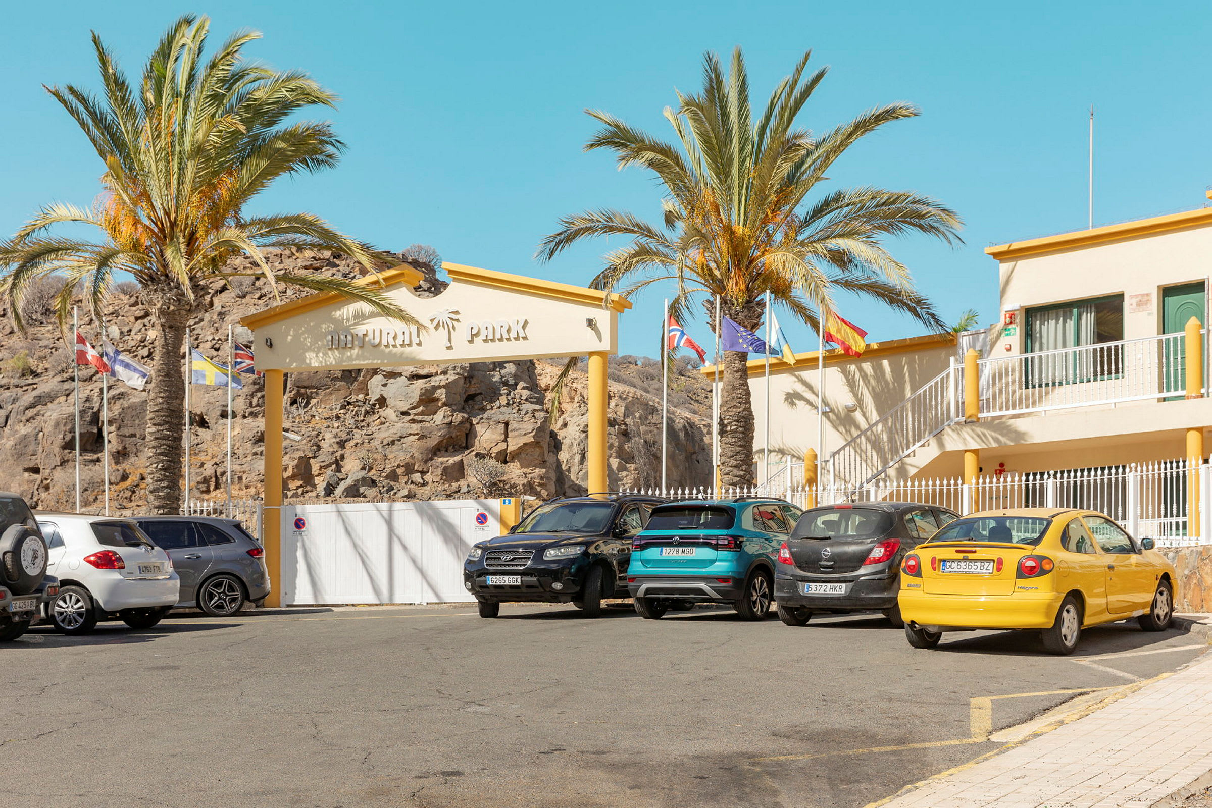Ingång till ett hotell med parkerade bilar och palmer i förgrunden. (AI-genererad text)