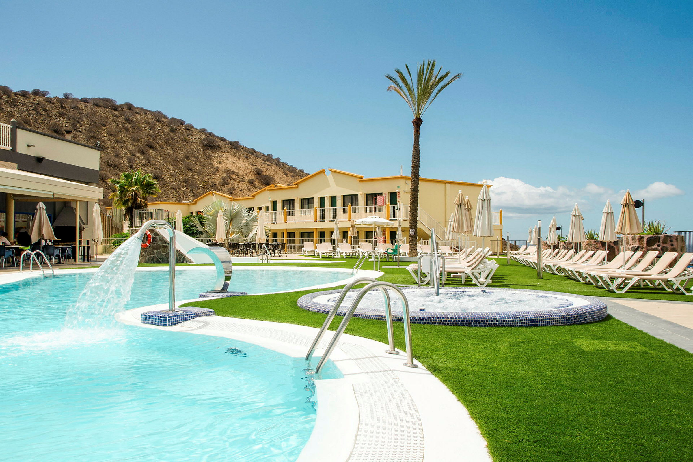 Ett hotell med swimmingpooler och solstolar, beläget nära ett berg. (AI-genererad text)
