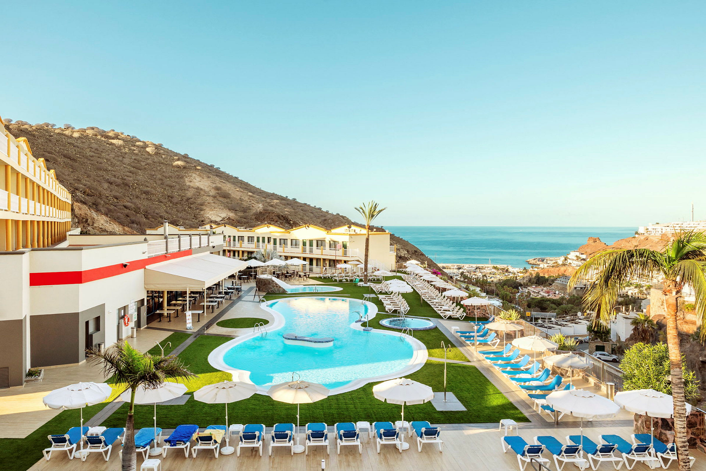 Ett semesterhotell med en pool, solstolar och parasoller, omgiven av en bergslandskap och utsikt över havet. (AI-genererad text)