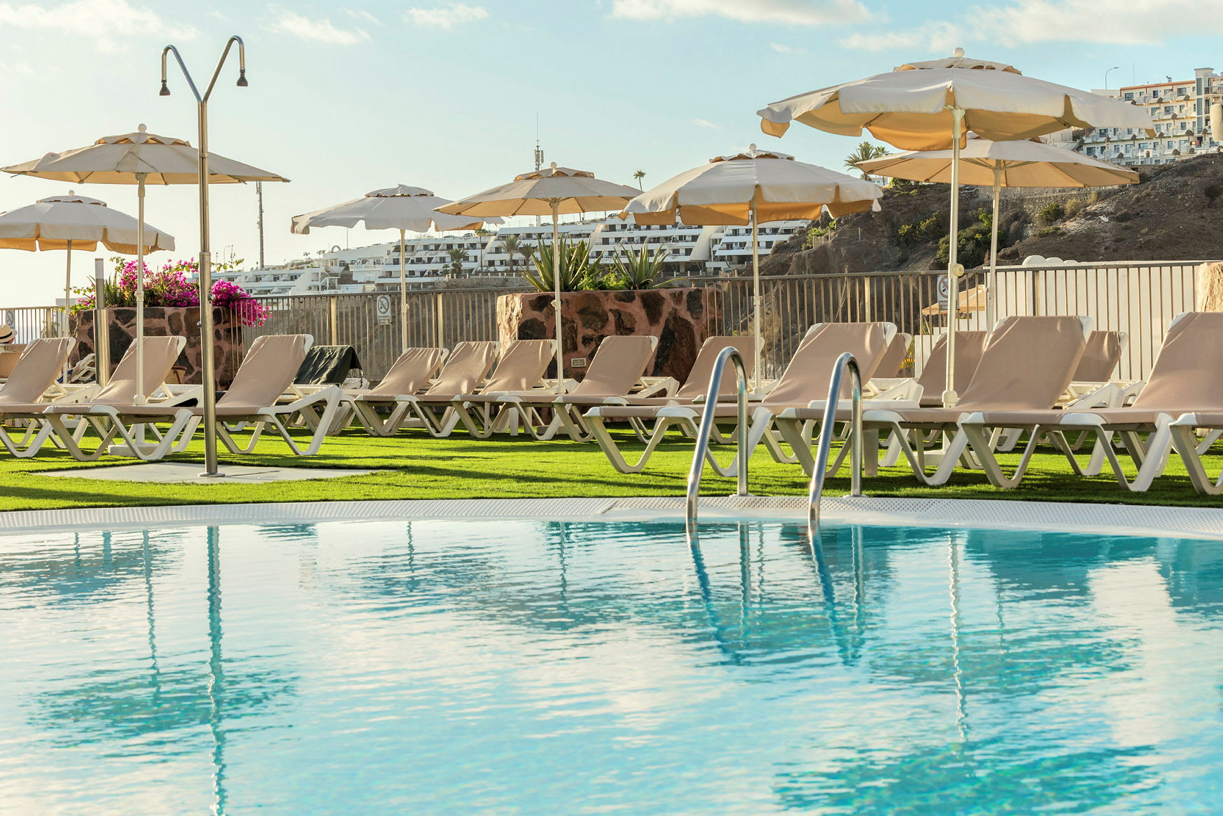 En pool med solstolar och parasoller runtomkring på en gräsmatta. (AI-genererad text)
