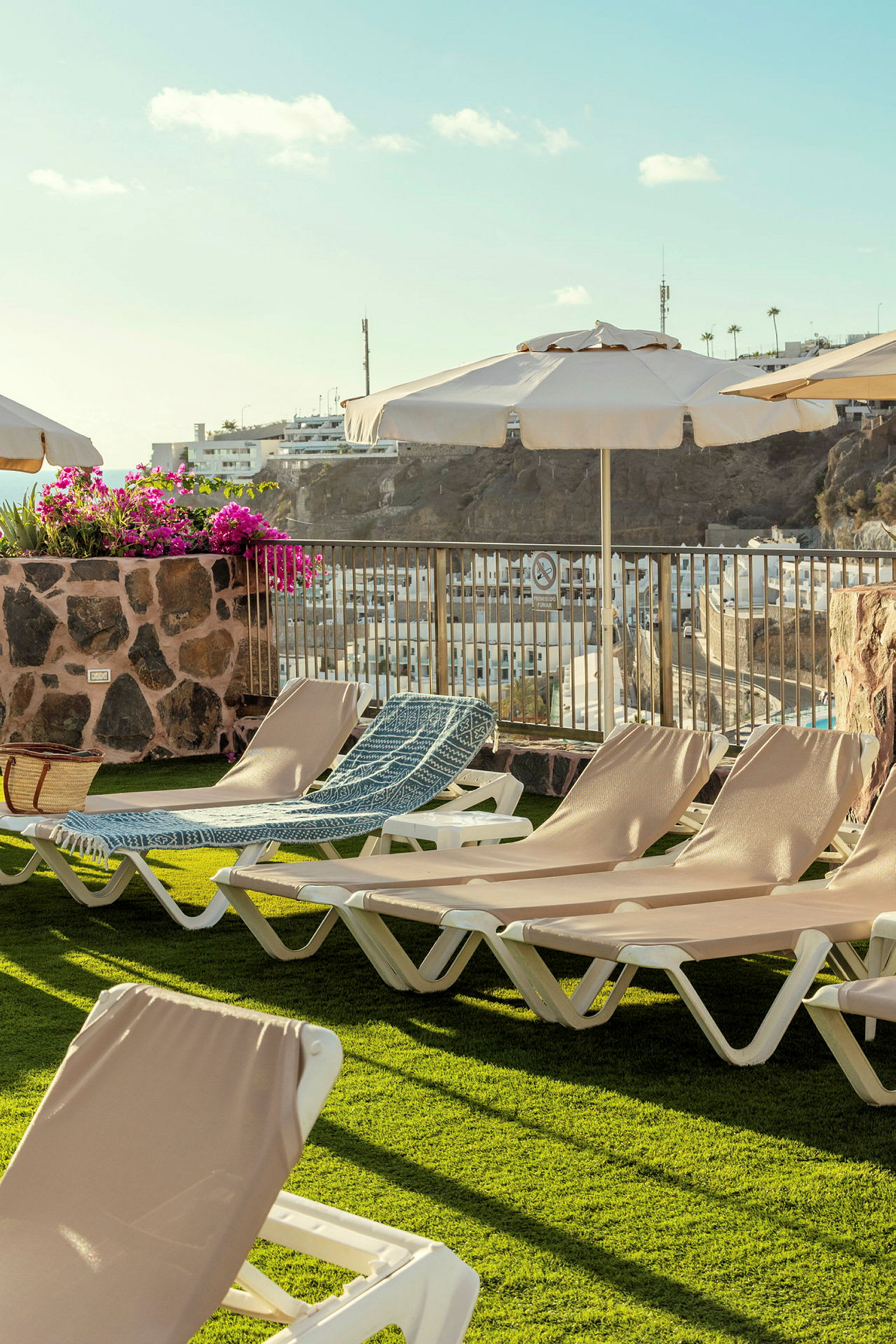 Bild av en solterrass med solstolar, parasoller och en blomsterkruka. I bakgrunden syns ett bostadsområde och kullar. (AI-genererad text)