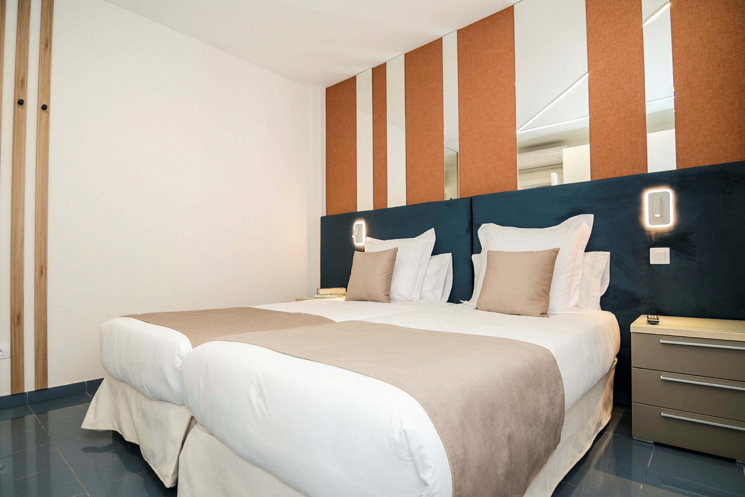 Ett modernt hotellrum med två enkelsängar bredvid varandra, bäddade med vita och beige sängkläder. Väggarna har speglar och orange paneler, och bredvid sängen finns ett nattduksbord. (AI-genererad text)