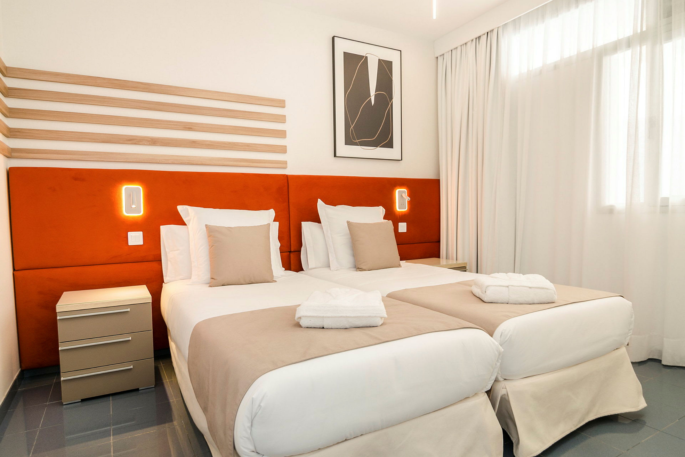 Ett hotellrum med två enkelsängar, orange sänggavlar, och vita sängkläder. En nattlampa syns på väggen. (AI-genererad text)