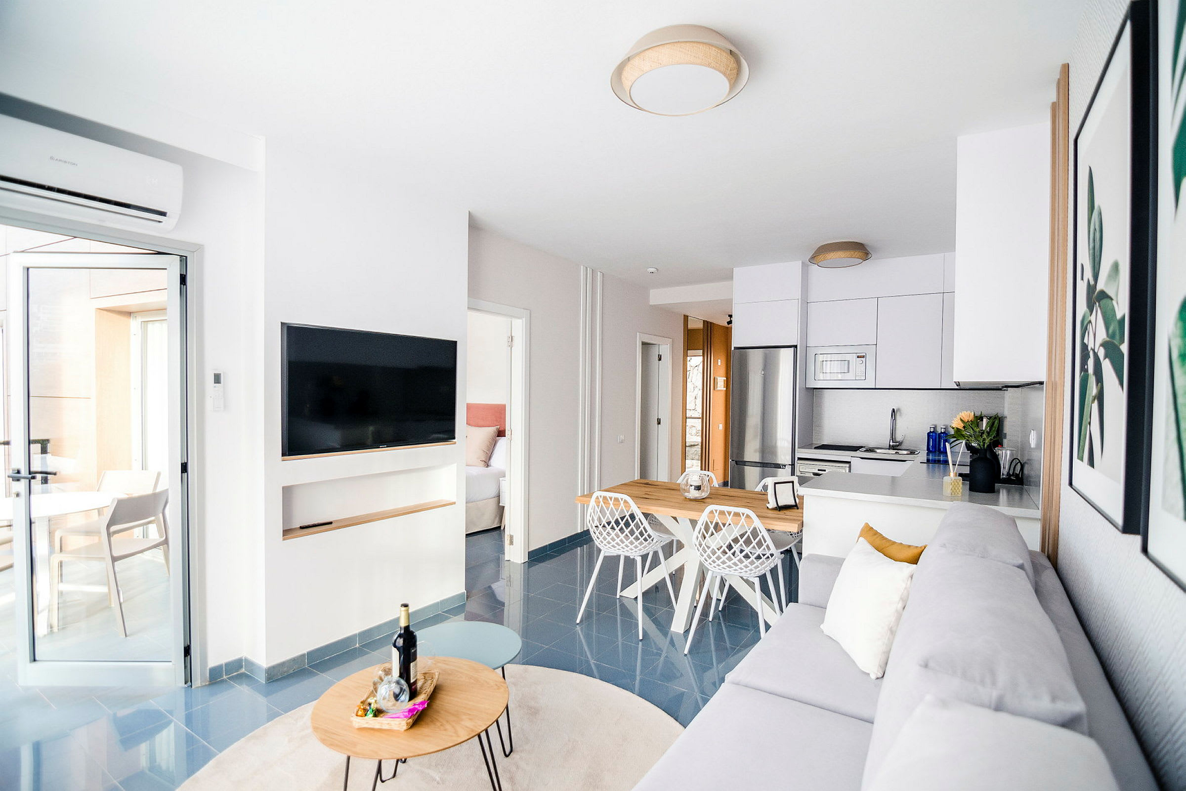 Ett modernt vardagsrum med öppen planlösning som inkluderar ett kök. Rummet har en grå soffa, en platt-TV på väggen och ett matbord med vita stolar. En dörr leder till ett sovrum och en annan ut till en ljus terrass. (AI-genererad text)