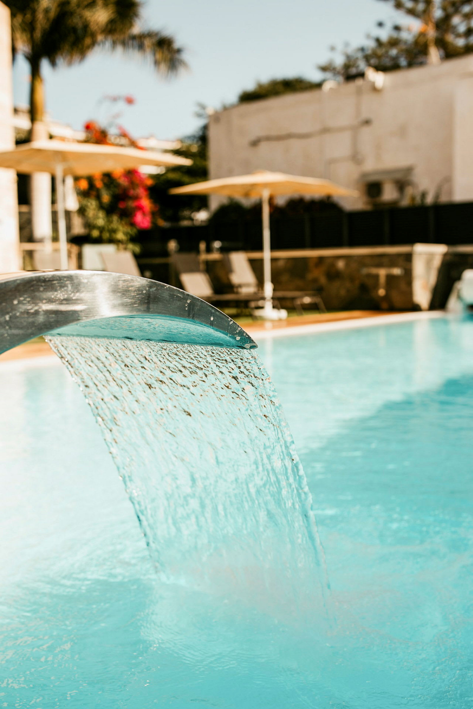 Närbild på en pool med vattensköld som rinner från en metallyta, med solstolar och parasoller i bakgrunden. (AI-genererad text)