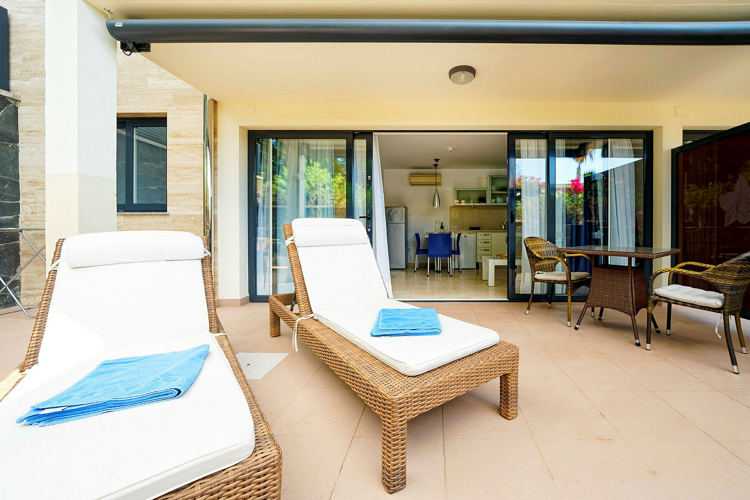 Två solstolar med vita dynor och en blå handduk på en terrass med bord och stolar framför glasdörrar till ett kök. (AI-genererad text)