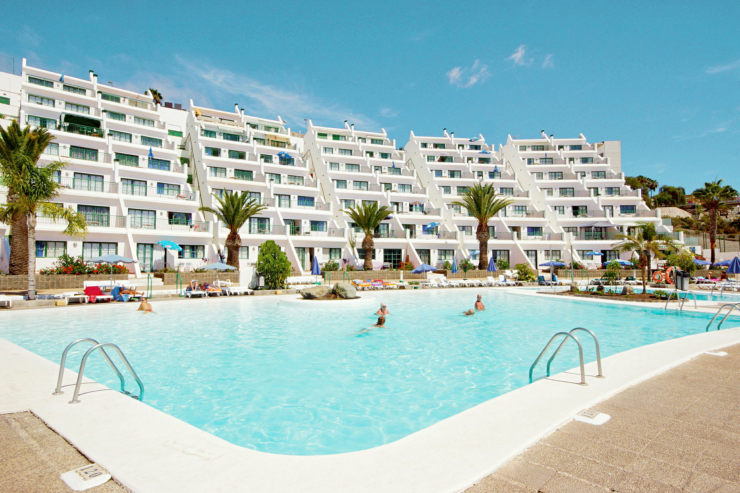 Hotellbyggnad med vit fasad och många balkonger, omgiven av palmer och en stor pool i förgrunden. (AI-genererad text)